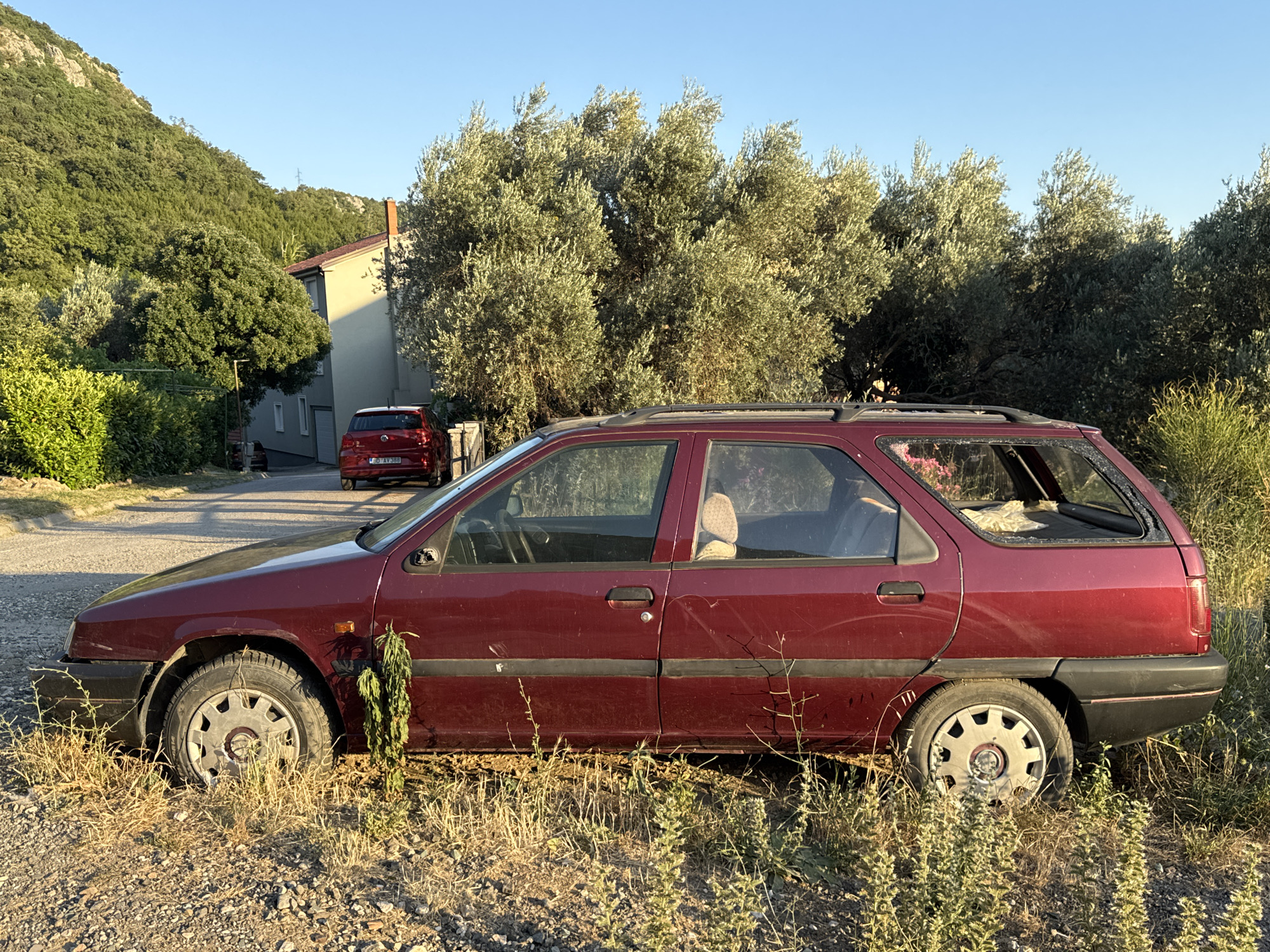 A maroon station wagon is parked on a dirt and gravel lot, surrounded by dry grass and sparse vegetation. The car is positioned sideways to the camera, with its front on the left and rear on the right. The vehicle appears to be older, with some visible wear and tear. In the background, a red hatchback is parked further down a paved road, near a light-colored building with a reddish-brown roof. Olive trees and other greenery form a dense backdrop, and a hillside covered in trees rises beyond the building. The sky is clear and blue, suggesting a sunny day. The location is the city of Rafailovići, Montenegro.