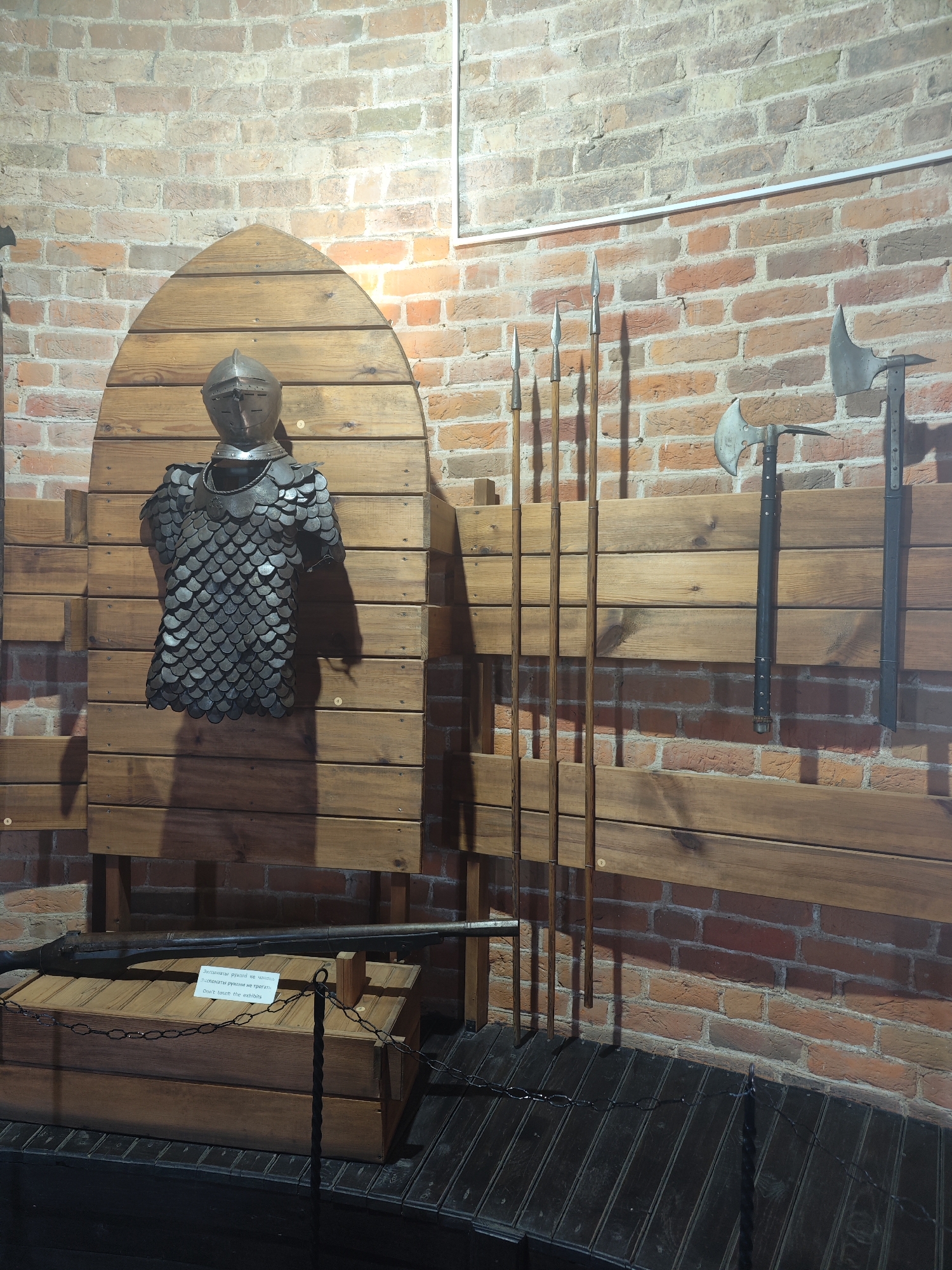 The image depicts a display of historical weaponry and armor inside what appears to be a museum or historical exhibit. The backdrop is a textured brick wall. In the center, a suit of scale armor with a helmet is mounted on a wooden display. To the right, several long spears are standing upright against the wall. Below the spears and to the far right, two axes are mounted on the wall. In the lower left corner, a long firearm, possibly a musket, rests on a wooden chest. A chain barrier is present in front of the firearm. The lighting suggests an indoor environment, likely with artificial lights casting shadows. The specific location within Kamyanyets, Belarus is not explicitly indicated by any visual cues in the image.