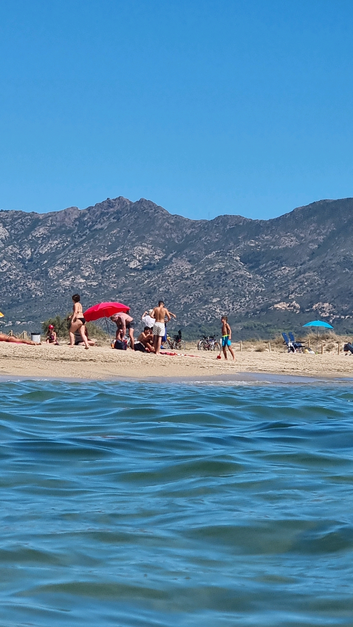 In Castelló d'Empúries, Spain, a sunny beach scene unfolds with a clear blue sky and a backdrop of rugged, sparsely vegetated mountains. The foreground is dominated by the rippling surface of the sea, rendered in various shades of blue. On the sandy shore, a group of people are engaged in various beach activities.
In the mid-ground, a vibrant red umbrella provides shade for several individuals. Some are seated, while others are standing. A man in white shorts and a white t-shirt is visible near the umbrella, as are other individuals in swim attire. Further to the right, a young boy in blue swim trunks stands on the wet sand at the water's edge, holding a red object, possibly a toy shovel. A bicycle with a basket is parked near the group, and another bicycle is visible further to the right. Two blue beach chairs and a blue umbrella are set up further down the beach, indicating other beachgoers. A woman in a black bikini walks away from the camera on the left side of the frame, and another person is lying on a towel further to the left. A small child in pink clothing is seated near the red umbrella. The sand is a light tan color, and the transition to the mountains is marked by dry vegetation.