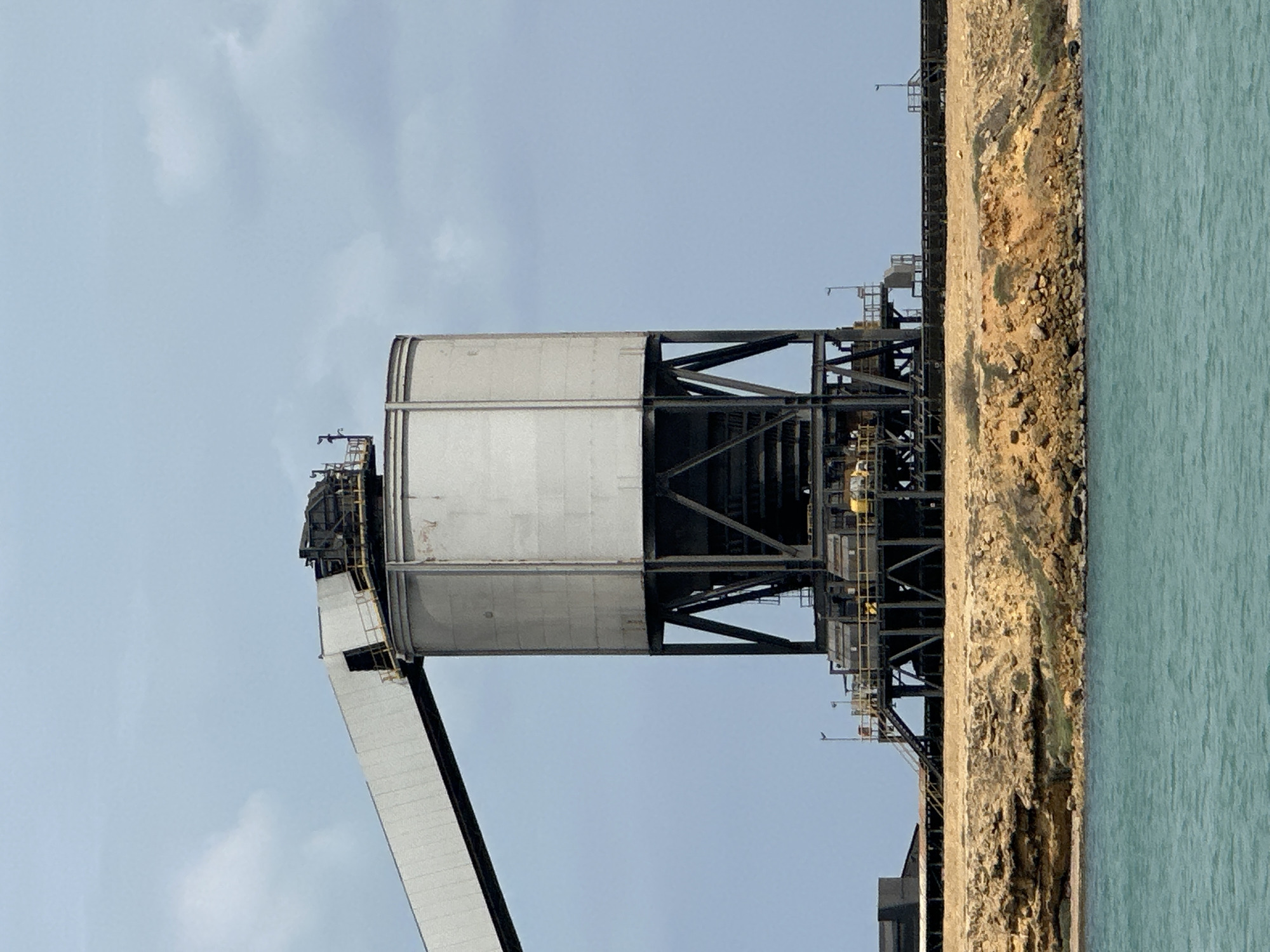 The image captures an industrial facility located by a body of turquoise water in Uribia, Colombia, under a light blue sky with sparse white clouds. A prominent feature is a large, light-grey cylindrical storage silo, elevated on a dark, multi-legged metal support structure. An enclosed, light-grey conveyor belt system extends from the upper left, connecting directly to the top of this silo, indicating material transport. Beneath the silo, a complex network of dark metal platforms, stairs, and railings is visible, with several small yellow pieces of equipment situated on the lower platforms. The facility rests on a rugged, eroded embankment of sandy-brown and orange rock that meets the water. An elevated dark pier or walkway extends horizontally to the right from the main industrial complex. No individuals are present in the scene.