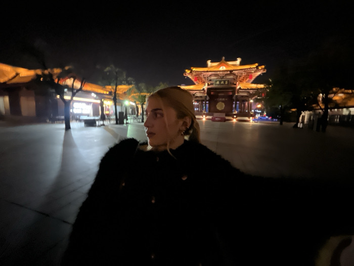 The image captures a nighttime scene within the Big Wild Goose Pagoda cultural area in Yanta District, Xi'an, China. In the mid-foreground, positioned slightly left of center, a person with blonde hair and fair skin is visible in profile, facing towards the right. This individual is wearing a dark, textured coat and an earring in their visible ear. The background features a large, ornate traditional Chinese gate structure with multiple tiered roofs, extensively illuminated by warm orange and yellow light, revealing Chinese characters on its facade. To the left and right of this prominent gate, additional traditional-style buildings with similar warm lighting line a wide, open paved plaza. Several indistinct figures are visible in the far background near these structures. The overall environment is dark, with artificial light sources illuminating the architectural elements and the ground, creating strong contrasts and subtle shadows.