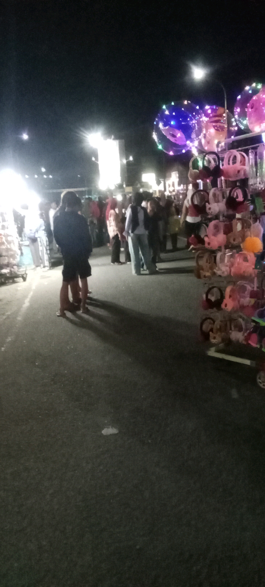 The image depicts a street scene at night, likely a night market, in the city of Pacitan, Indonesia. Several individuals are visible, appearing to be shoppers or vendors. In the foreground, two figures stand with their backs to the camera, one wearing dark clothing and shorts, the other in darker attire. To the right, a stall displays an assortment of goods, including colorful stuffed animals and small bags, suspended on racks. Above the stall, illuminated balloons with embedded lights float in the dark sky. Further back, a crowd of people mingles, with the glow of streetlights and market stalls illuminating the area. The ground is paved, suggesting a street or public walkway. The overall atmosphere is one of a lively evening gathering or market.