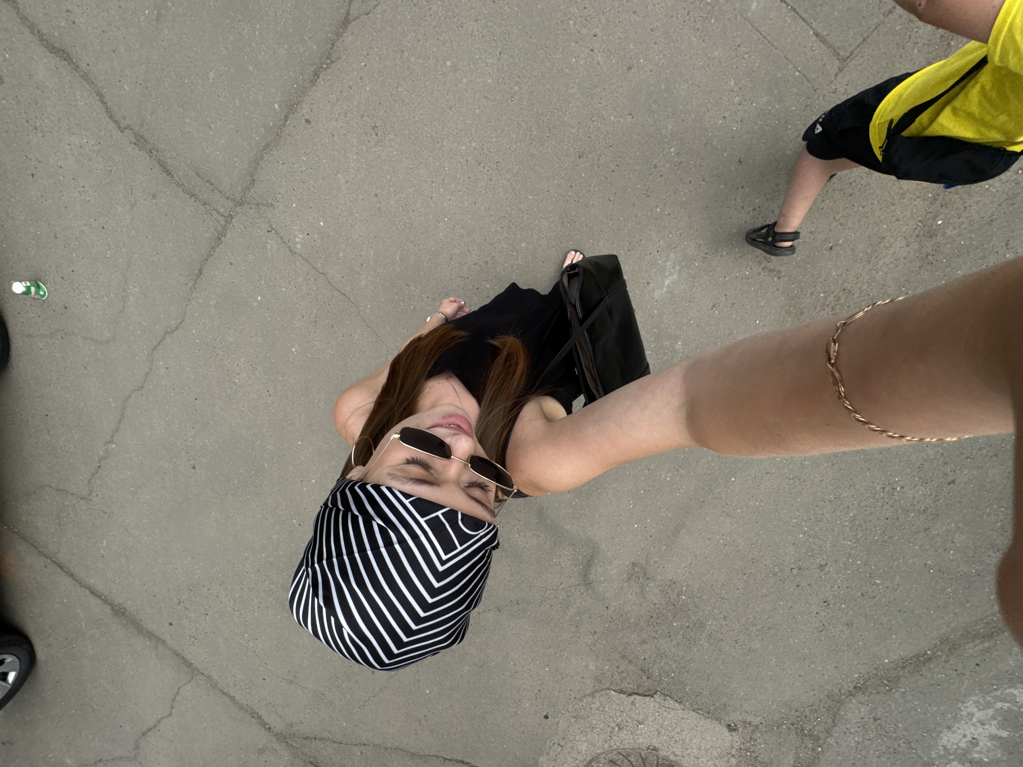This media file shows two people from a high-angle perspective on a cracked asphalt surface, possibly a street or pathway, under bright daylight. The overall setting appears to be outdoors, with a slight overcast sky suggested by the lighting.
The primary subject is a young woman wearing sunglasses, a black dress, and a black and white striped bandana or headscarf. She is holding a black bag and has a delicate gold bracelet on her arm. She is looking upwards and to the side, with a slight smile, giving an impression of a casual and carefree moment.
In the background, another person is partially visible, wearing a bright yellow shirt and black shorts, with black sandals. This person is walking away from the camera.
A small green and white object, possibly a can or bottle, is visible on the ground to the left. The concrete surface is worn and shows signs of age with numerous cracks.
The scene suggests a casual outing, possibly during the daytime with fair weather. The location context provided is Chaikovsky, Russia, although no specific landmarks are visible to confirm this. No text is discernible in the image.