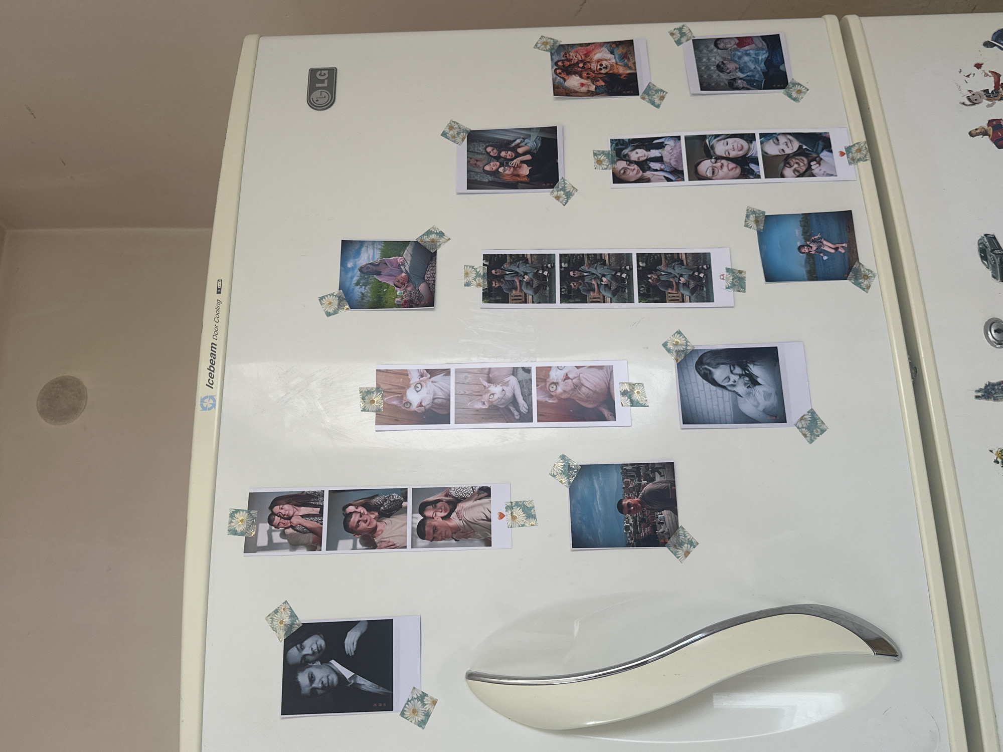 The image shows the side of a white LG refrigerator with various photographs and stickers attached to it. The photographs appear to be of people and pets, with some appearing to be polaroid-style prints. The stickers are small and feature a daisy pattern. The refrigerator is in a room with light-colored walls. The image is taken from a slightly elevated angle, looking down at the refrigerator. The city of Gyumri, Armenia is not mentioned or depicted in the image.