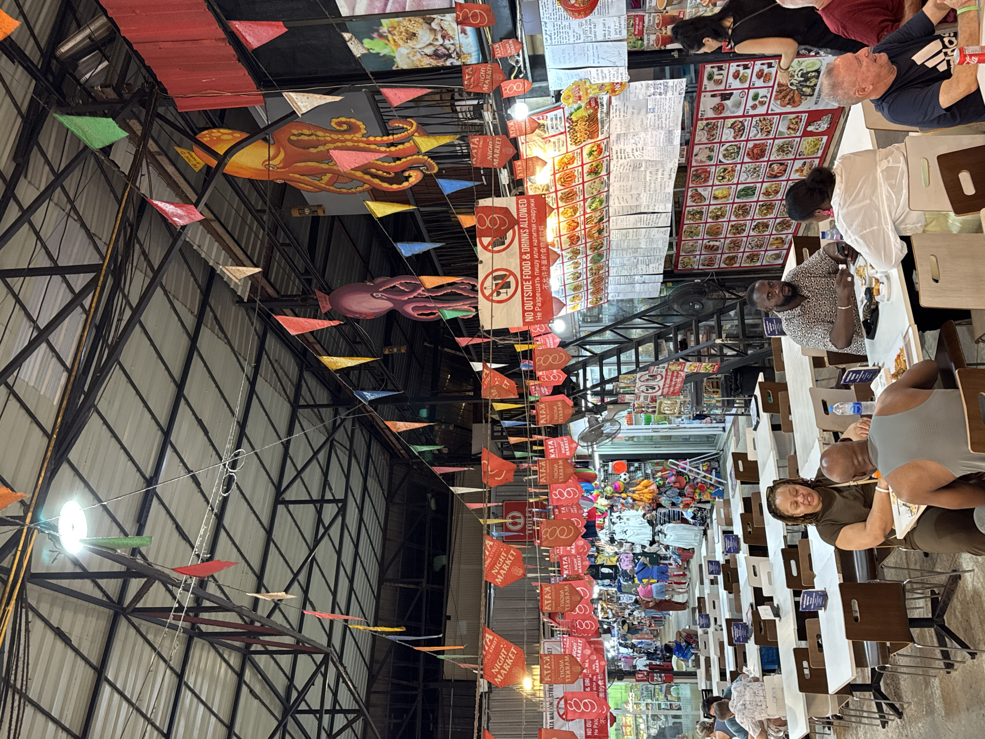 This image depicts a vibrant night market scene in Karon, Thailand. The setting is a covered outdoor market with rows of food stalls and shops. The overhead structure is made of metal beams and corrugated metal roofing. Colorful triangular flags and decorative octopuses are strung across the market, adding to the festive atmosphere.
Several people are visible, engaged in various activities. Some are seated at tables, enjoying meals, while others appear to be browsing or working in the stalls. The mood seems lively and bustling, characteristic of a busy night market. The lighting suggests it is evening or nighttime, with artificial lights illuminating the area.
Visible text includes signage for "KATA NIGHT MARKET" and "ATA WALKING STREET". There is also a prominent sign stating "NO OUTSIDE FOOD & DRINKS ALLOWED" in English, Russian, and Chinese, indicating a diverse clientele. Other signs appear to be menus with food images and prices. The overall impression is a lively, commercial hub with a focus on food and local crafts.