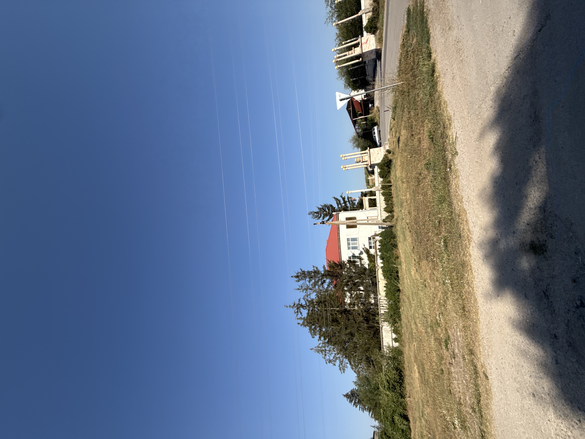 The image depicts a clear, bright day with a vast expanse of blue sky dominating the upper portion. Three thin, white power lines stretch horizontally across the sky. In the mid-ground, a white building with a red roof is visible, surrounded by mature green trees and dense foliage. A low, light-colored fence encloses the property. To the right of the building, an ornate gateway with multiple columns and decorative tops stands by the roadside. A silver car is parked near this gateway. Further down the road, another similar gateway and more trees are present. The foreground consists of a dirt and asphalt road with patches of dry grass on the left side. Shadows from unseen objects, possibly trees, are cast across the road in the lower right corner of the frame. The scene appears to be in a semi-rural or suburban setting within the city of Kuibyshevskoye rural settlement, Ukraine.