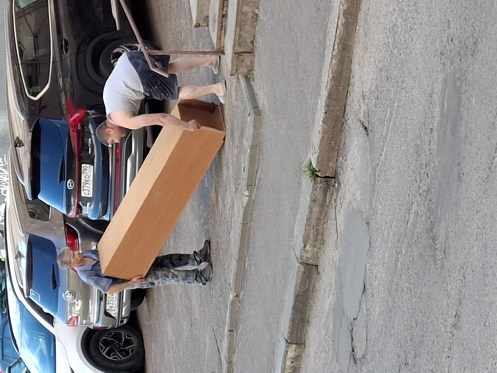 Two men are carrying a large, flat, wooden object, possibly a large panel or door, down an asphalt street. The man on the left is wearing a blue short-sleeved shirt and camouflage pants, and is holding the object with both hands. The man on the right is wearing a grey tank top and dark shorts, and is bent over, holding the object with his left hand. He is wearing light-colored slip-on shoes. The man on the left is wearing dark sneakers. Several cars are parked on the right side of the street behind them, including a white SUV, a grey SUV, and a black SUV. The black SUV has a license plate that reads "C379KO 790". A set of concrete stairs is visible on the far right of the image. The street is cracked and uneven, with some patches of green vegetation growing through the cracks.