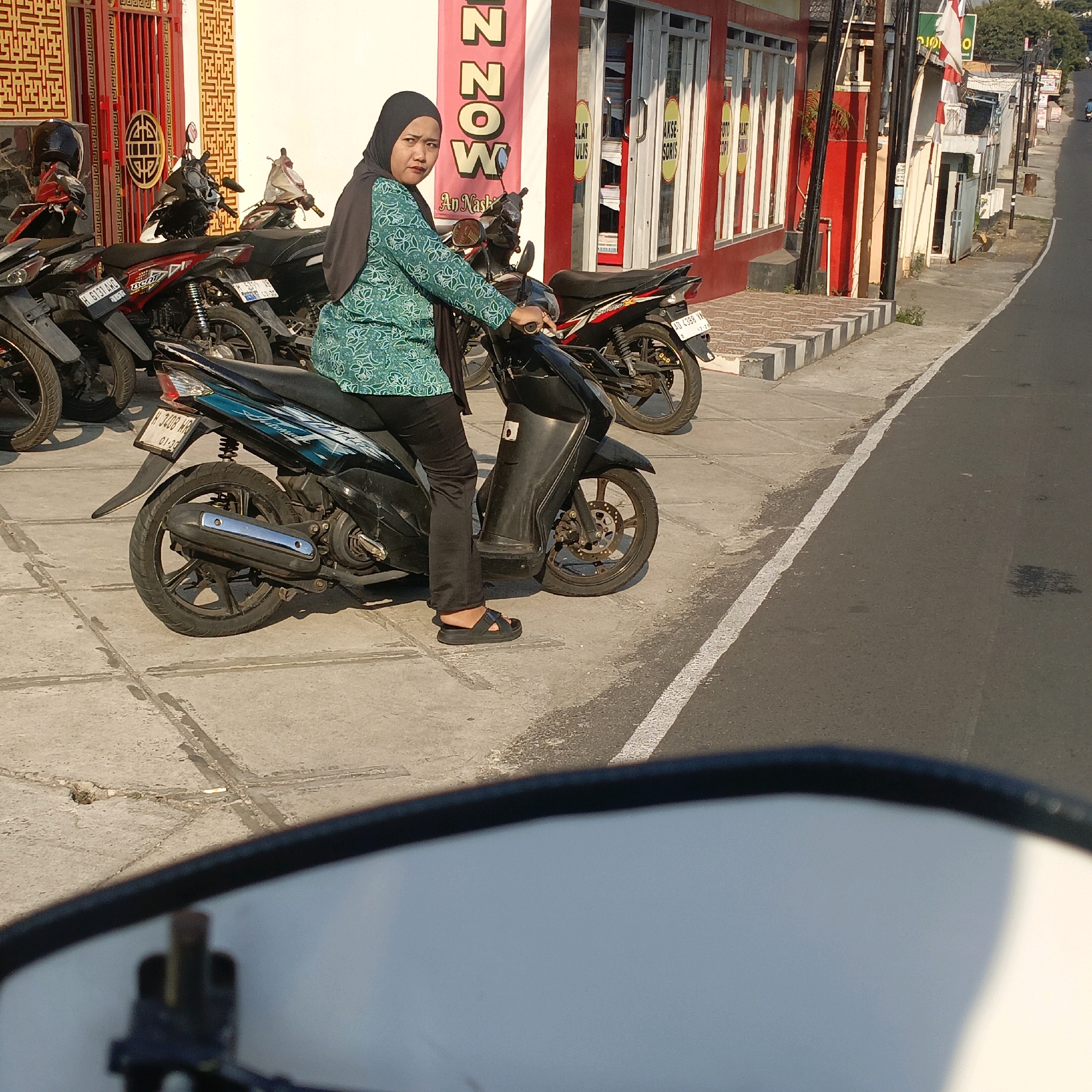 A woman wearing a black hijab and a green patterned tunic is seated on a black scooter. She is looking towards the camera with a neutral expression. The scooter is parked on a paved sidewalk in front of a building. Several other motorcycles are parked behind her. The building has red accents and windows displaying various items. A sign with red lettering that reads "NOW" is visible on the building. The scene appears to be in a commercial area, likely in the city of Salatiga, Indonesia, given the presence of Indonesian license plates on the motorcycles and the architectural style. The sun is casting long shadows, suggesting it is late afternoon or early morning. The sidewalk is made of concrete tiles, and a road with a white line marking runs alongside it.