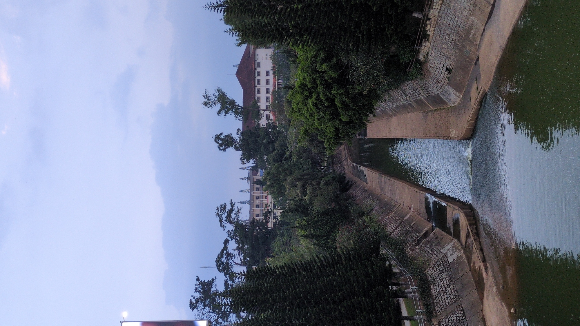 The image shows a dam and reservoir in Da Lat, Vietnam. The structure is made of stone and concrete. Water flows over a series of stepped weirs. The water appears green and still in the reservoir. Lush vegetation, including tall pine trees, surrounds the dam. Two buildings are visible in the distance: a large white structure with a red roof and a smaller building with multiple spires. The sky is overcast.