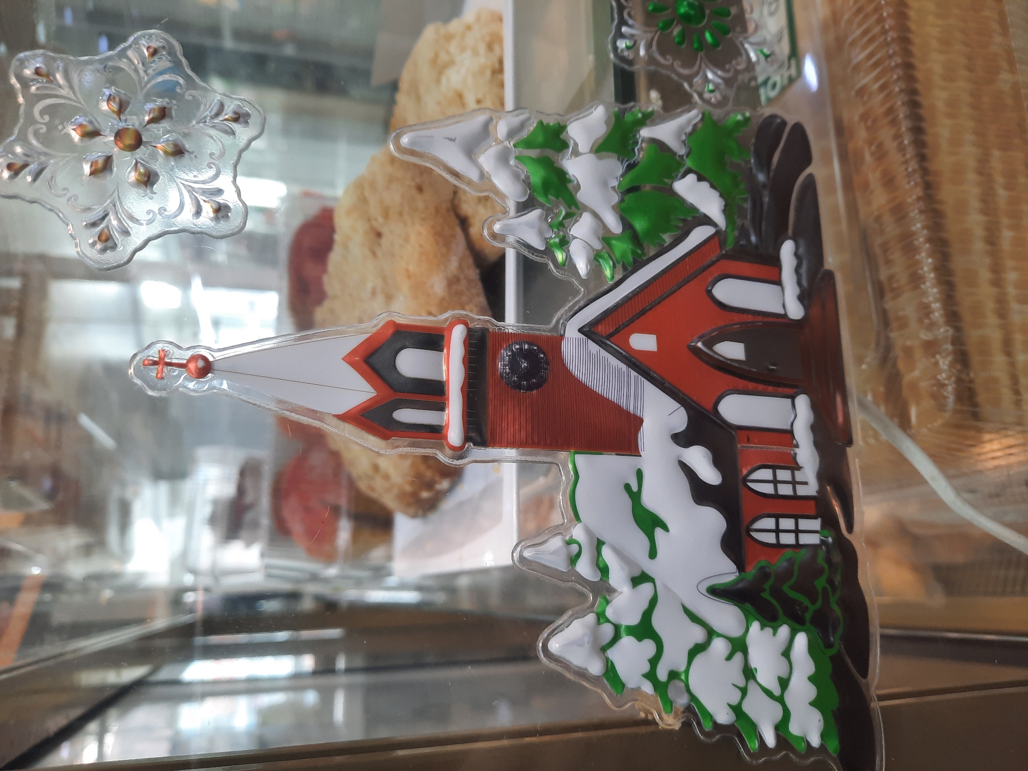 The image displays a close-up view of decorative items arranged within a glass display case, likely in a commercial establishment such as a bakery or cafe. The central subject is a clear acrylic ornament depicting a church in a snowy landscape. The church features a distinct red roof, white walls, and a tall white steeple topped with a small red cross. Black accents outline windows, the belfry, and architectural details. Surrounding the church are evergreen trees, rendered in green with white snow details, creating a winter scene.
To the upper left, another clear, textured decorative item shaped like a snowflake or star is visible. It has an ornate pattern with a central brown/gold embellishment and similar smaller accents.
In the background, out of focus, are various baked goods, appearing golden brown and somewhat textured, possibly pastries or bread. The clear glass of the display case creates reflections of interior lights and blurred elements. A white power cord is visible along the right edge of the frame. The overall scene suggests a festive, possibly holiday-themed, display in a food-related business. No legible text is present in the image.