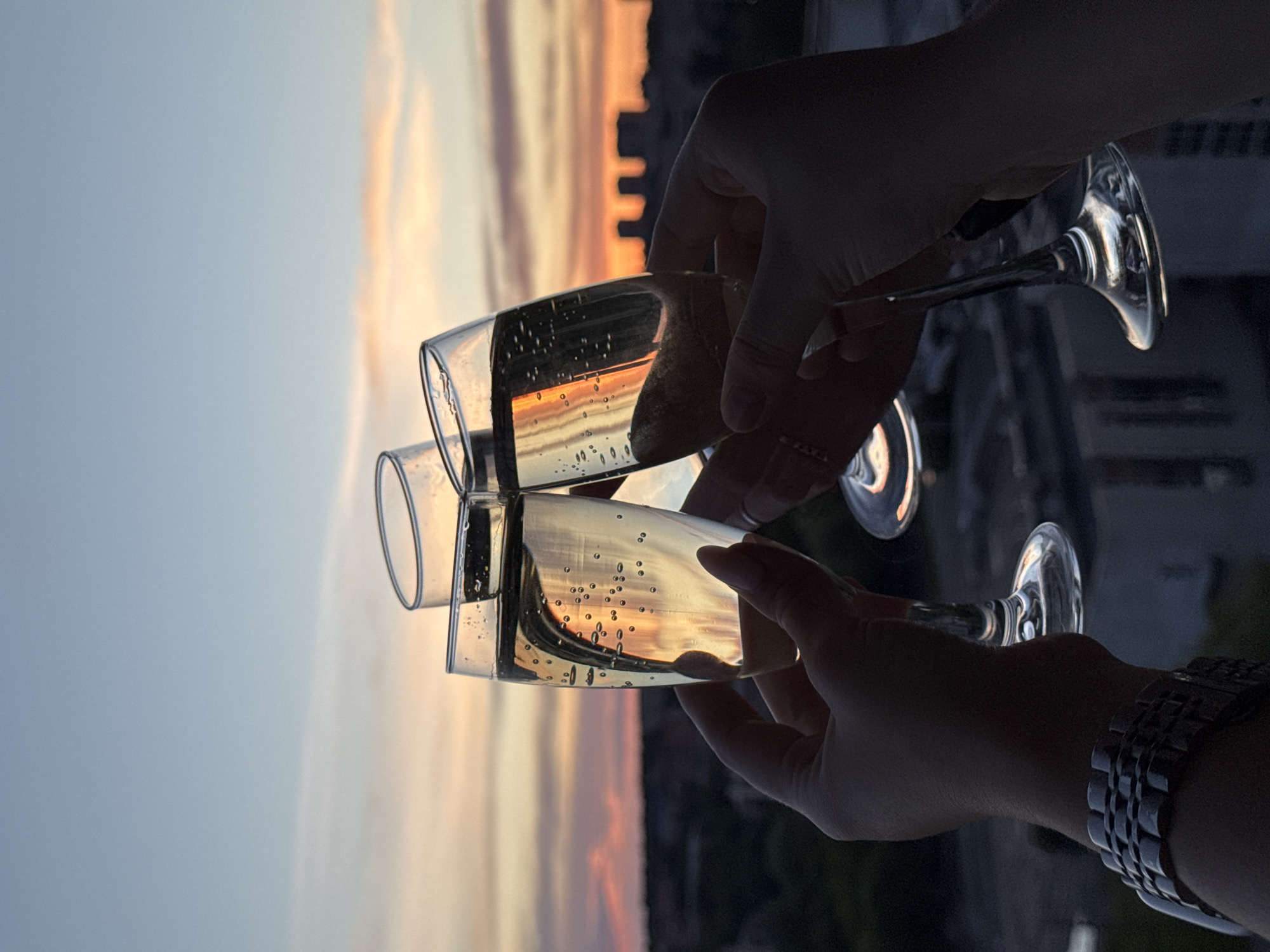Two hands, one wearing a silver watch, are holding two champagne flutes in front of a sunset. The sunset reflects in the champagne flutes. The location is unidentified and there is no indication this is in Izhevsk, Russia.