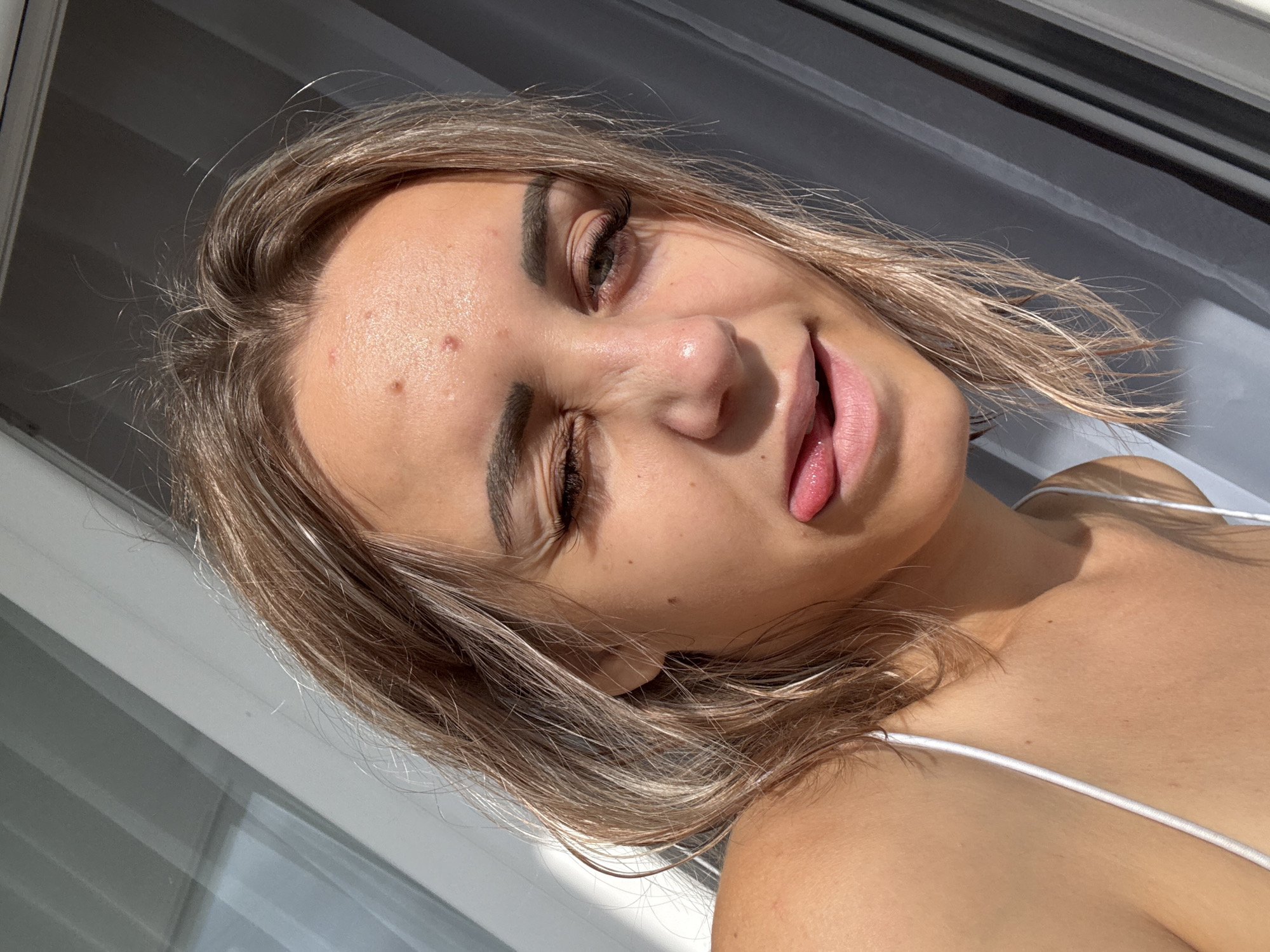 In the city of Kholmsk, Russia, a young woman with blonde, shoulder-length hair is captured in a selfie. She is looking towards the camera with her left eye squinted and her tongue sticking out slightly. The natural sunlight illuminates her face, highlighting her skin texture and the presence of a few blemishes on her forehead. She is wearing a white strap, suggesting she is dressed in a top or swimwear. The background consists of a window with vertical blinds, casting diagonal shadows across the scene. The overall impression is a casual, playful moment captured outdoors.