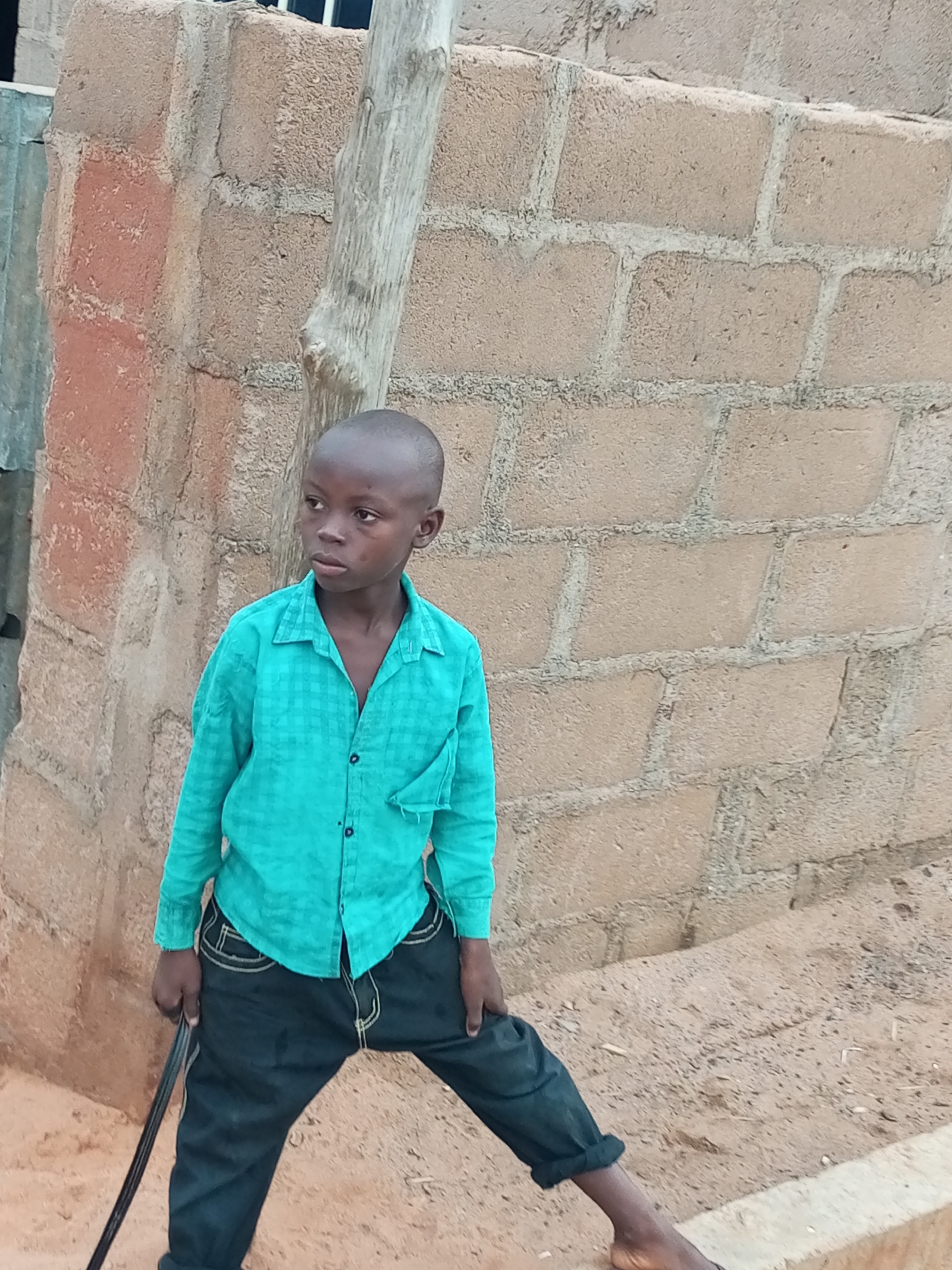 A young boy with a shaved head stands outside in what appears to be a village or rural area. He is looking off to his left, with a slightly serious expression. He is wearing a bright teal button-down shirt with a checkered pattern and dark blue jeans. His left hand rests on a black object that is partially visible, possibly a stick or cane. The background is a rough, textured wall made of light-colored bricks. To the left of the boy, a metal fence is visible. The ground is uneven and dusty. The overall impression is of a casual outdoor scene in a non-urban setting.