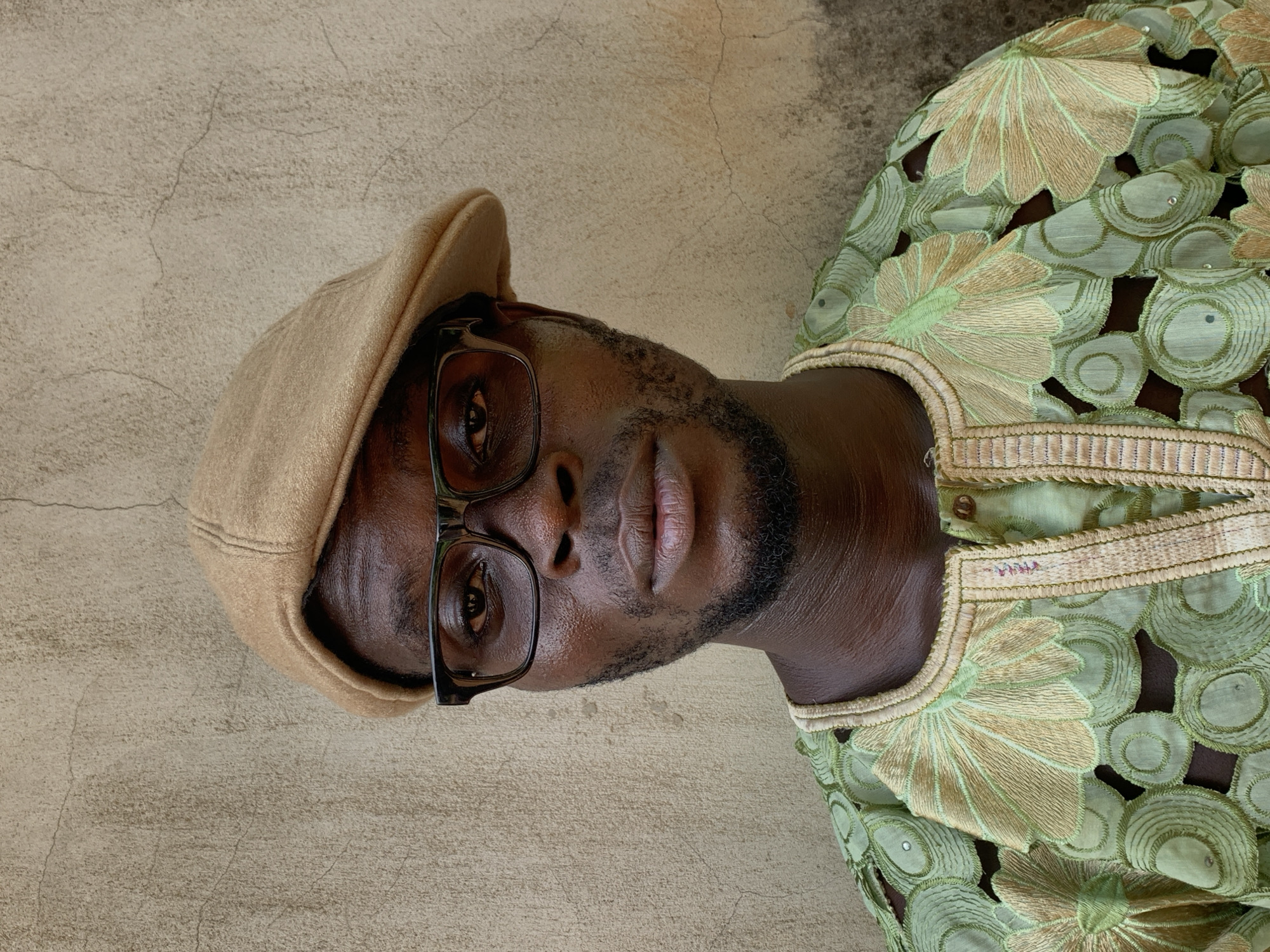 A medium close-up portrait of a man is presented, rotated 90 degrees counter-clockwise. The man, who has dark skin, is looking directly at the camera with a neutral expression. He wears a light tan or beige flat cap and dark-framed rectangular glasses. A neatly trimmed beard or stubble is visible on his chin and jawline.
He is dressed in what appears to be traditional Nigerian attire, likely an Agbada or similar garment, which is predominantly light green. The clothing features elaborate embroidery in a combination of lighter green and gold/tan hues, forming large floral and circular patterns. Some sections of the fabric appear to be cut out, and small, reflective embellishments (possibly sequins or rhinestones) are scattered within the embroidered patterns. The neckline and trim of the garment are intricately bordered with woven gold and tan threads.
The man is positioned against a plain, textured wall that is light beige or off-white in color. The wall shows subtle signs of age and wear, with fine cracks visible across its surface. The lighting is natural and even, suggesting the photograph was taken outdoors during the daytime. There is no visible text in the image. The setting is consistent with the provided location context of Ikorodu, Nigeria.