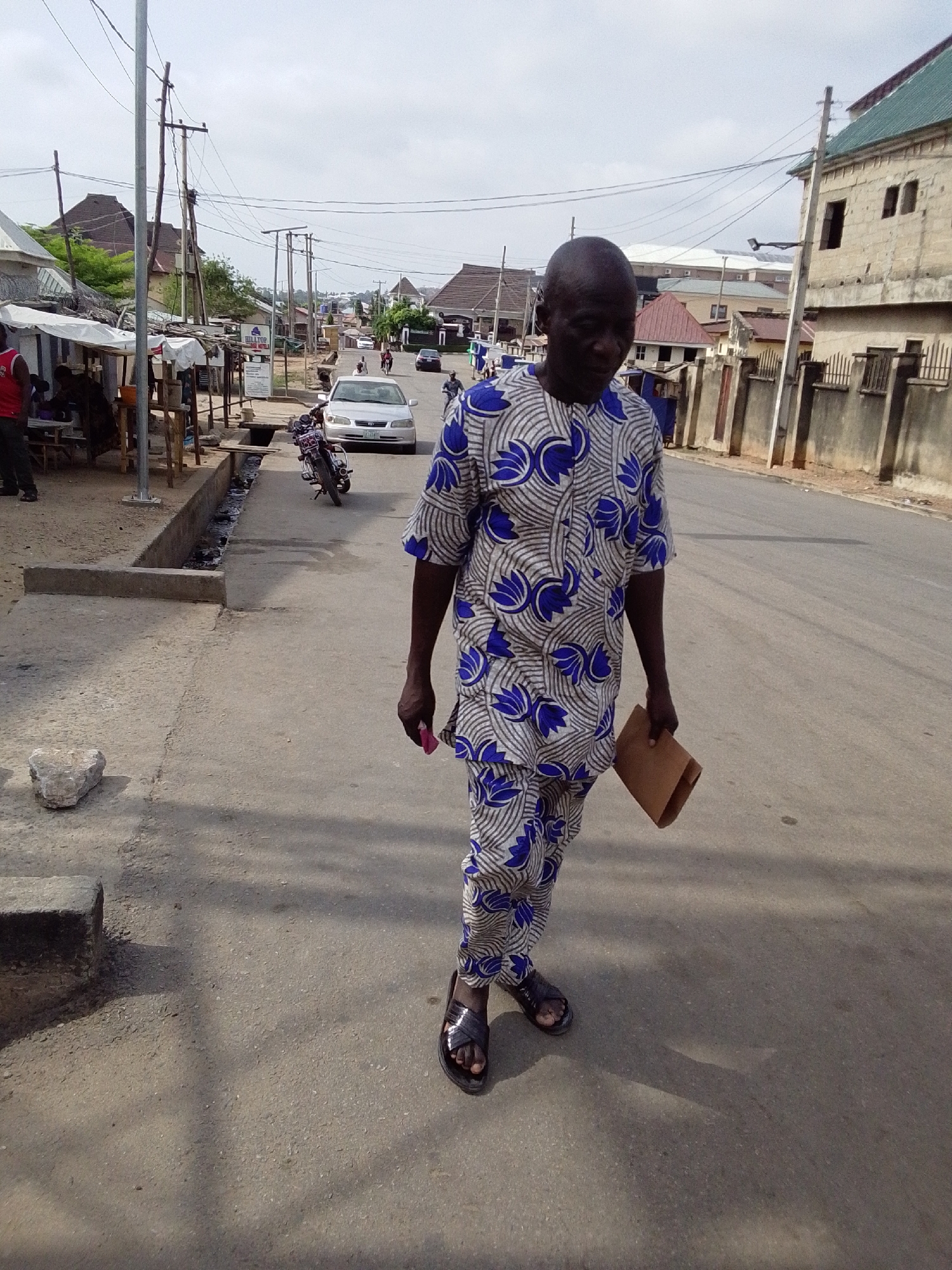 The image depicts a man walking on a paved street in Lokoja, Nigeria, under an overcast sky during the daytime.
The main subject is a dark-skinned, bald man of average build, seen from the waist up in the foreground. He is wearing a two-piece traditional Nigerian outfit with a distinctive blue, white, and grey pattern, featuring stylized floral or wave motifs. He has dark sandals on his feet. In his right hand, he holds a small, folded brown paper bag or envelope, and in his left hand, a small pink object, possibly a phone. His gaze is directed downwards.
The street is made of asphalt, with concrete curbs and an open drainage ditch running alongside the left edge. A large light-colored rock rests on the ground near the ditch. On the left side of the road, several makeshift market stalls with simple wooden frames and tarp covers are visible, with people partially obscured behind them. A white sign near these stalls shows the text "HILLTOP... HOTELS".
Further down the street, a silver sedan (possibly a Toyota) is parked, and a motorcycle is positioned nearby. In the background, various buildings line the street, including a multi-story concrete structure with bare walls and a green-tiled roof on the right. Numerous wooden utility poles with a dense network of overhead electrical wires crisscross the sky, characteristic of the urban infrastructure. The overall scene suggests a bustling, semi-urban environment.