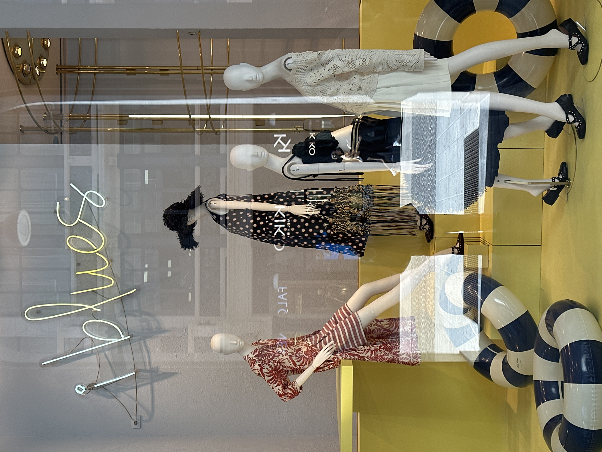 This is a vertical photograph of a store window display in Milan, Italy. The display features multiple mannequins dressed in fashion items, arranged in a layered presentation.
The subjects include at least four mannequins, each displaying a different outfit. The mannequins are positioned at different heights and angles, with some leaning.
Objects in the display include clothing, shoes, and decorative elements. One mannequin wears a white lace top over a white dress. Another is dressed in a black top and skirt ensemble, with the word "KIKO" visible on the skirt. A third mannequin wears a black and white polka dot dress with a fringed hem. The bottom mannequin is adorned in a red and white striped top and a patterned skirt. Several black shoes with decorative studs are visible on the mannequins. In the background, there are striped inflatable rings, resembling life preservers, and a yellow wall. A neon sign with cursive lettering, possibly spelling "Shop," is visible on the left side of the window.
The scene appears to be a retail setting, specifically a fashion boutique. The display is designed to showcase the clothing and create an appealing visual presentation for shoppers. The lighting suggests it is daytime, possibly late afternoon or evening given the reflections on the glass.
Notable details include the fashion styles of the clothing, the specific brand name "KIKO" visible on one outfit, and the neon sign. The overall aesthetic of the display is modern and stylish, typical of a fashion-forward retail environment. The reflection on the glass shows part of the street outside, hinting at the location being a public thoroughfare.