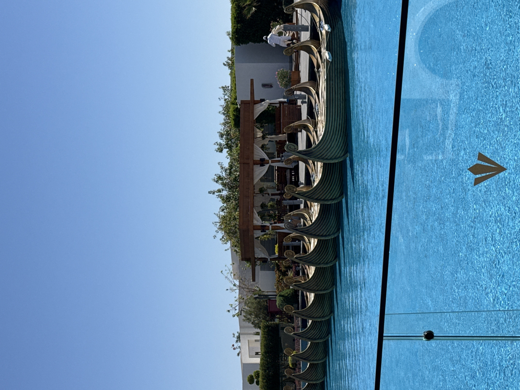 The image depicts an outdoor scene featuring a swimming pool with bright blue water. Along the edge of the pool, a series of decorative, boat-shaped objects are lined up. These objects have a bronze or gold metallic finish and feature intricate designs, resembling ancient ships with ornate prows. One person, dressed in white, is visible near the boats, possibly attending to them or engaged in an activity.
Behind the pool, there is a landscaped area with lush green bushes, trees, and some architectural elements. A wooden structure with white canopies suggests a seating or relaxation area. The background includes modern buildings with white walls. The sky is a clear, bright blue, indicating a sunny day with good weather. The overall setting appears to be a resort or a hotel in a warm climate, consistent with the context of Sharm El Sheikh, Egypt.
The image is captured from a high angle, looking down and across the scene. The sunlight creates reflections on the water's surface and highlights the metallic details of the decorative boats. The style of the boats and the resort-like environment suggest a tourist destination focused on leisure and relaxation. No visible text is present in the image.