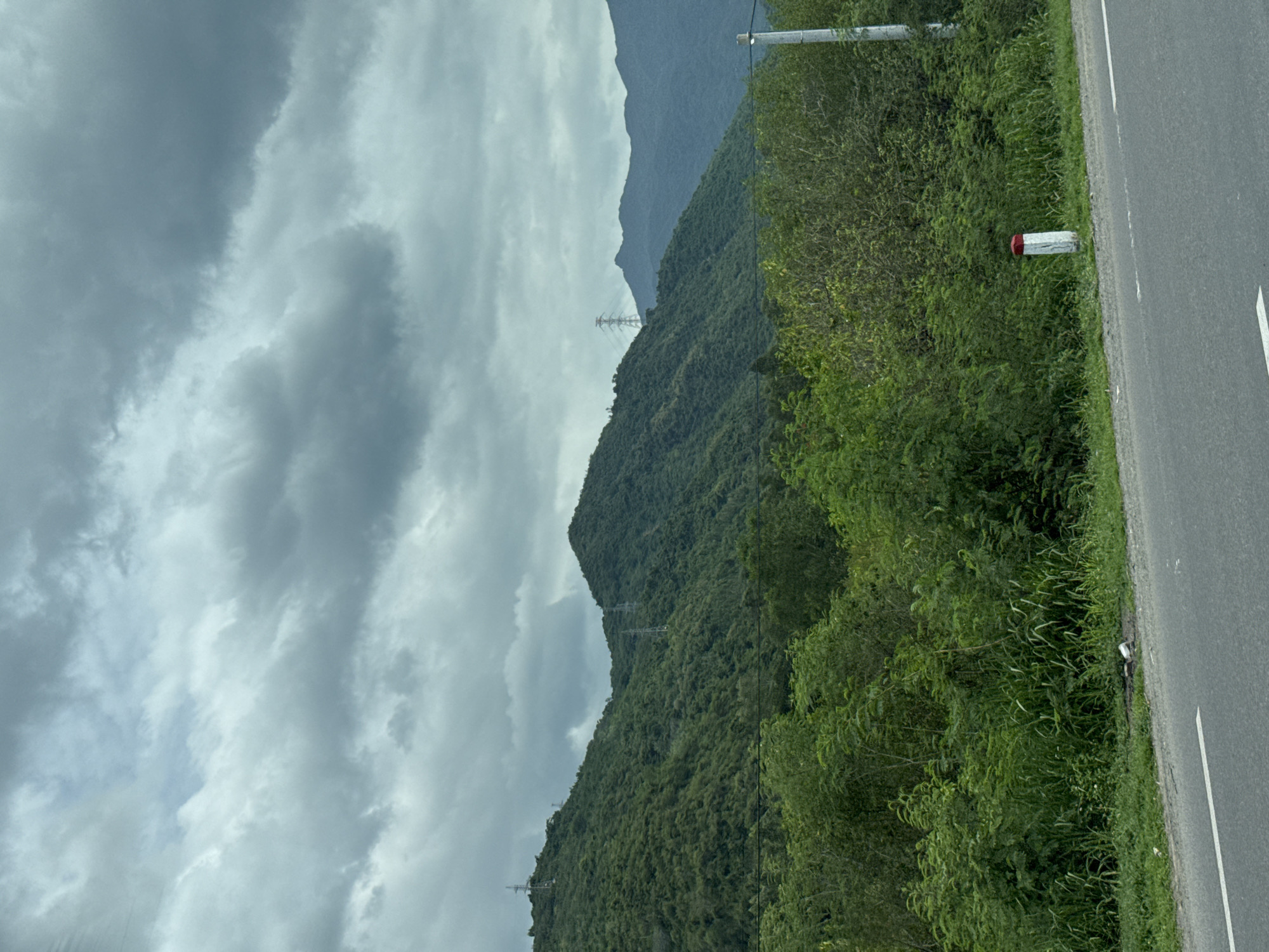The image depicts a scenic view of lush green mountains under a dramatic, cloudy sky, likely taken from the side of a road in Nam Ninh Hòa, Vietnam. The foreground features a grey asphalt road with a white lane marker, bordering a dense area of green vegetation. This vegetation includes tall grasses and bushes, with some taller trees visible further back. A white post with a red cap stands near the roadside. In the mid-ground, the rolling hills are covered in thick, green foliage, with several tall transmission towers dotted across their slopes, indicating the presence of electrical infrastructure. The background reveals more distant, hazy mountain ranges under a heavily overcast sky with dark, textured clouds. The overall impression is one of a rural, natural landscape characterized by its mountainous terrain and dense greenery, with man-made structures subtly integrated into the environment.