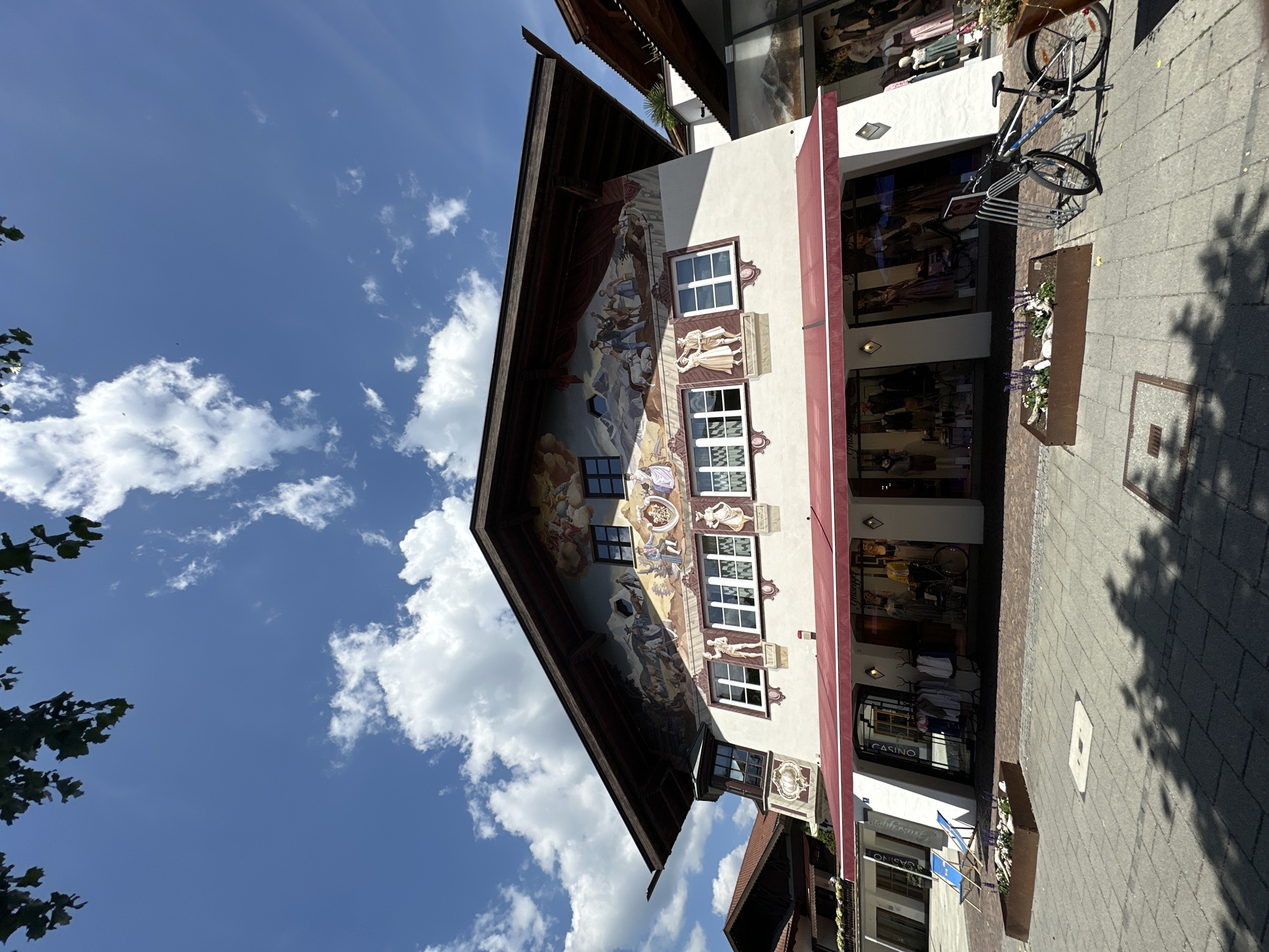 The image shows a building in Garmisch-Partenkirchen, Germany, with a mural depicting figures and scenes from Bavarian culture. The building has a red awning over its storefronts, which display clothing and mannequins. A bicycle is parked in front of the building, and two blue chairs are placed on the cobblestone sidewalk. The sky is blue with white clouds, and tree branches are visible in the upper left corner.