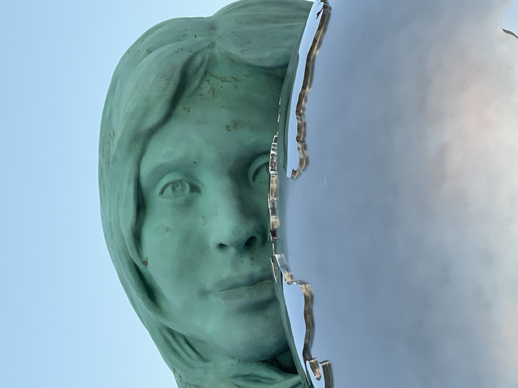 A close-up, low-angle shot captures a section of a large, weathered, teal-green statue of a woman's head, made of bronze or a similar material. Her face is tilted downward, with her right eye partially visible and her mouth slightly open. The statue rests on a reflective, metallic sphere that appears to be a globe, with an outline of landmasses etched into its surface. The background is a clear, pale blue sky. The statue is located at the "Eternal Flame" memorial on Victory Square in Krasnodar, Russia.