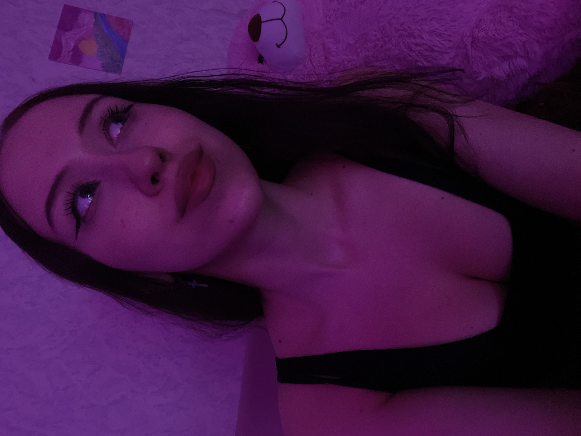 A young woman with dark hair, wearing a black top, is shown from the chest up. She has on eye makeup and lipstick. She is looking upwards and to her right. To her left, her shoulder and upper chest are visible. To her right, a large, pink, fuzzy teddy bear is partially visible. Behind her, a wall with textured wallpaper is illuminated by purple lighting. A small painting is hanging on the wall. The scene is indoors, likely in a residential setting. There is no information in the image that places the scene in Votkinsk, Russia.
