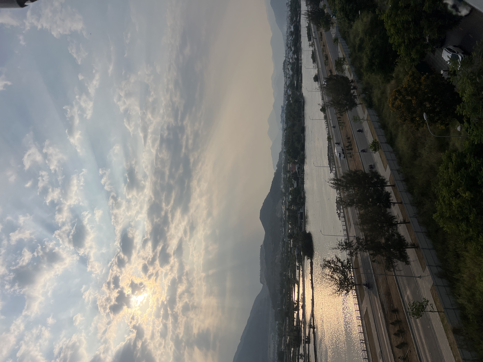 The image shows a landscape with a river, mountains, and city elements. The sky is filled with clouds, and the sun shines through them. The river reflects sunlight. A road runs along the river's edge, with trees and some buildings. The mountains are visible in the background. The location is in or near Nam Natural Heritage Area Trang, Vietnam.