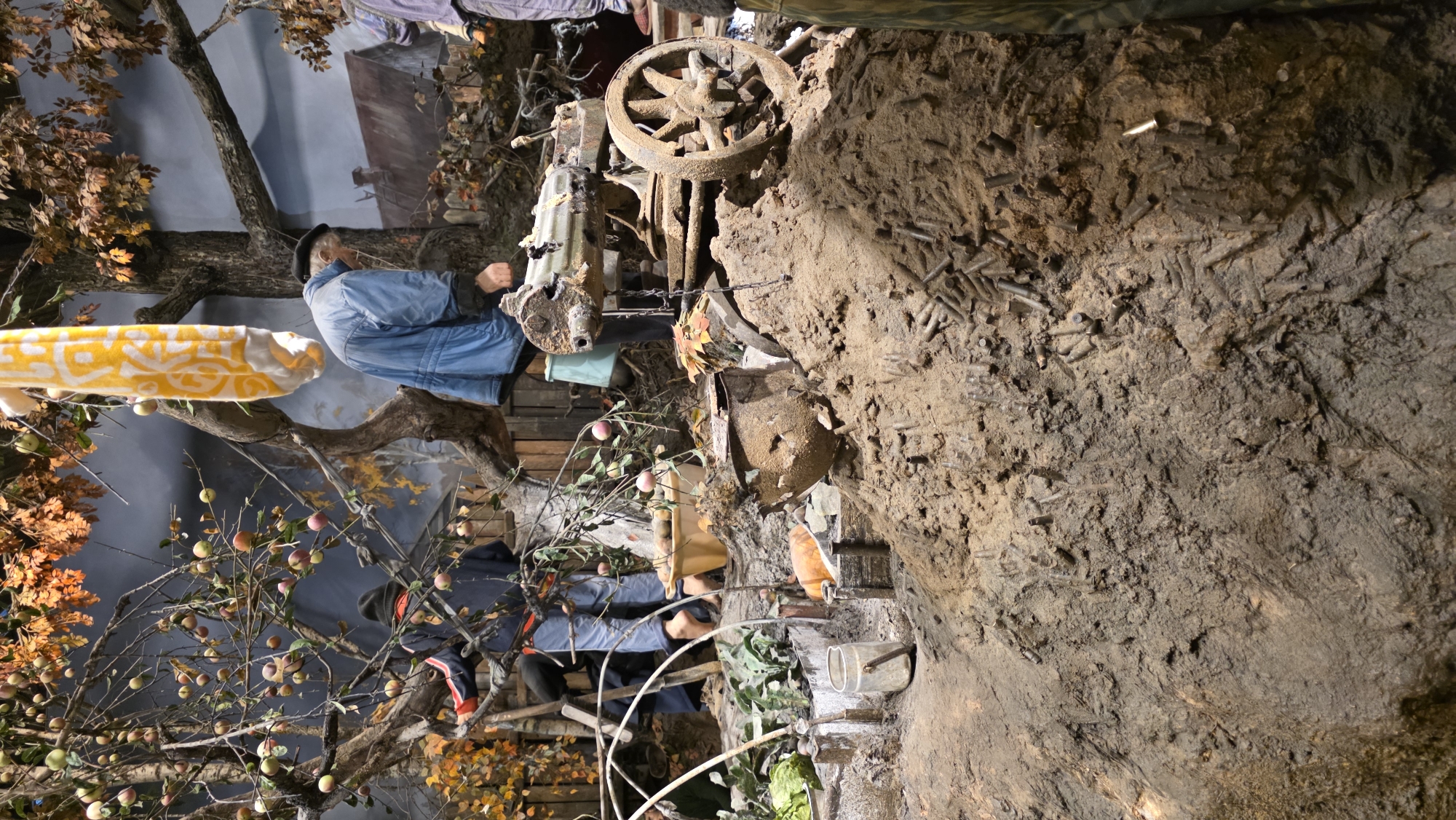 This image depicts a diorama, likely from a museum exhibit. The scene is set outdoors, featuring a tree laden with apples, a figure wearing a blue jacket and cap seemingly operating an old-fashioned machine, and other figures wearing jackets. A dirt or mud wall is seen with small, cylindrical objects embedded, possibly representing ammunition. The background includes painted elements mimicking landscapes and trees. The location is not specified in the image, denoted only as Unknown, Unknown.