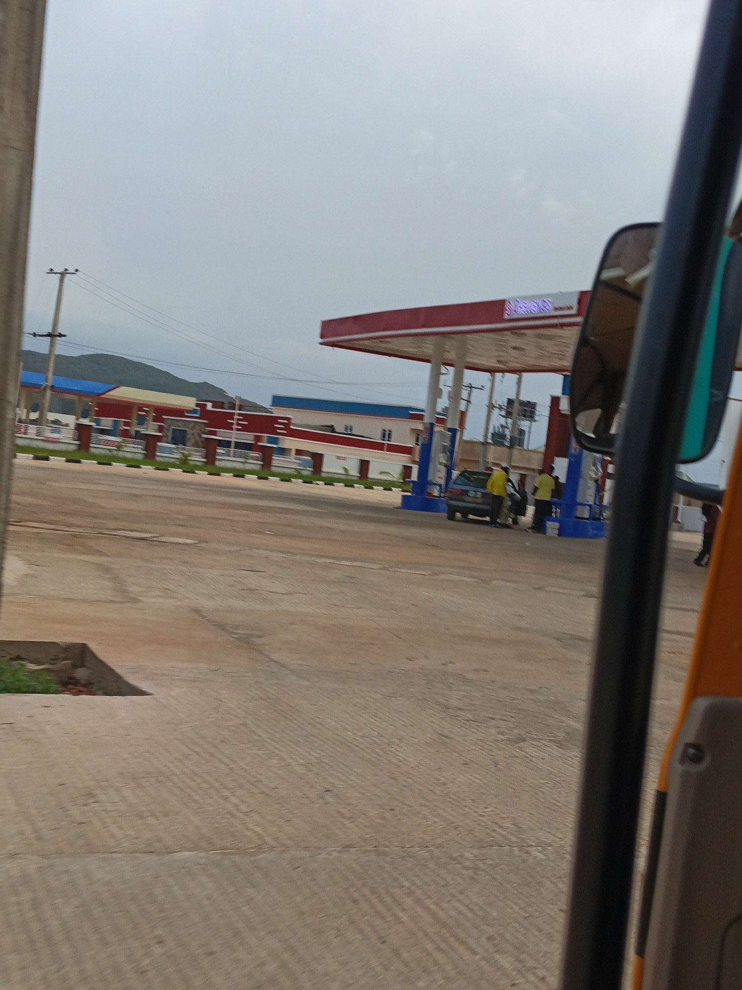 The image shows a gas station with red and white canopies in Kuru Dungus, Nigeria. A dark sedan is parked at one of the pumps, with at least three people, two wearing yellow shirts, interacting with it. The gas station has a prominent red and white sign with blue lettering. In the background, there are buildings with blue and red accents, and further in the distance, hills are visible under an overcast sky. The ground is paved with concrete, showing a textured pattern. The foreground is dominated by the interior of a vehicle, with a black pole and part of an orange and gray structure visible. Power lines and poles are also present.