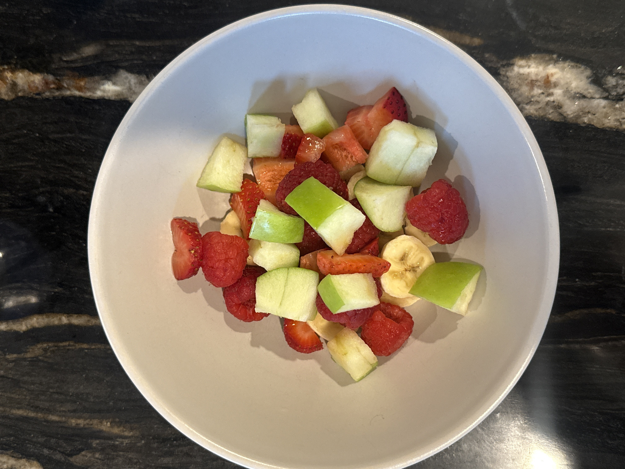A bowl of chopped fruit sits on a black and white granite countertop. The bowl is white, and the fruit mix includes diced green apples, raspberries, strawberries, and banana slices. The location appears to be a domestic kitchen, potentially within Southlake, United States of America.