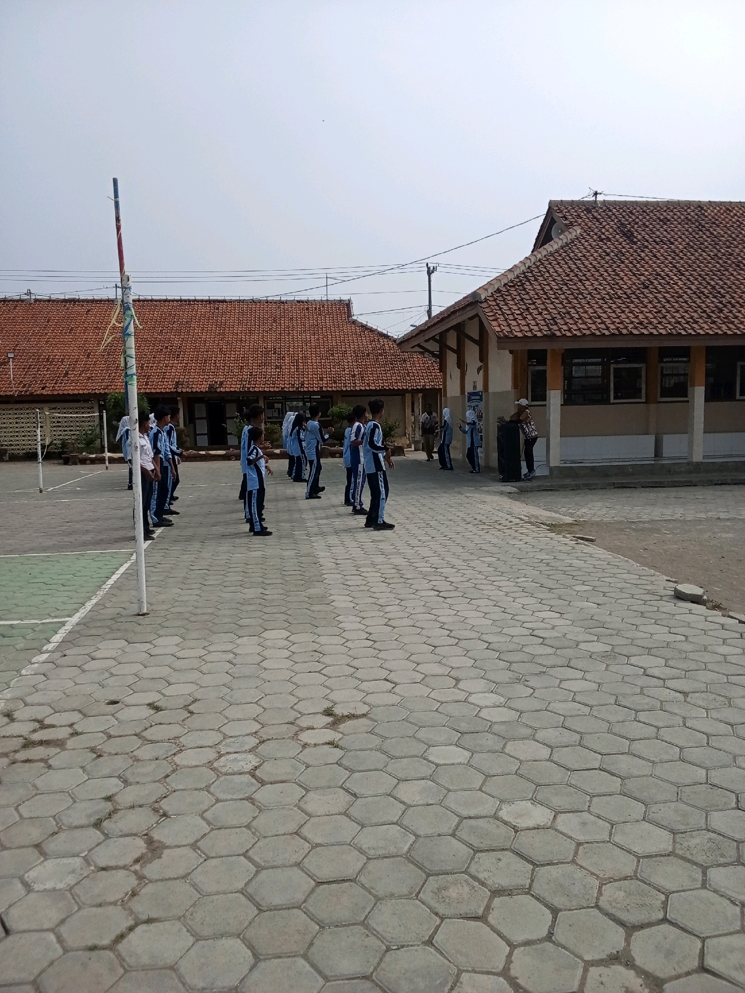 A group of students in light blue uniforms stand in formation on a paved hexagonal tile courtyard. They appear to be participating in an outdoor assembly or drill. Behind them are two buildings with terracotta tile roofs and open-sided shelters. A tall pole with white and red bands stands to the left of the frame, likely a flagpole. The courtyard is well-maintained with some patches of grass growing between the tiles. The scene suggests a school or community gathering in an urban or semi-urban setting.