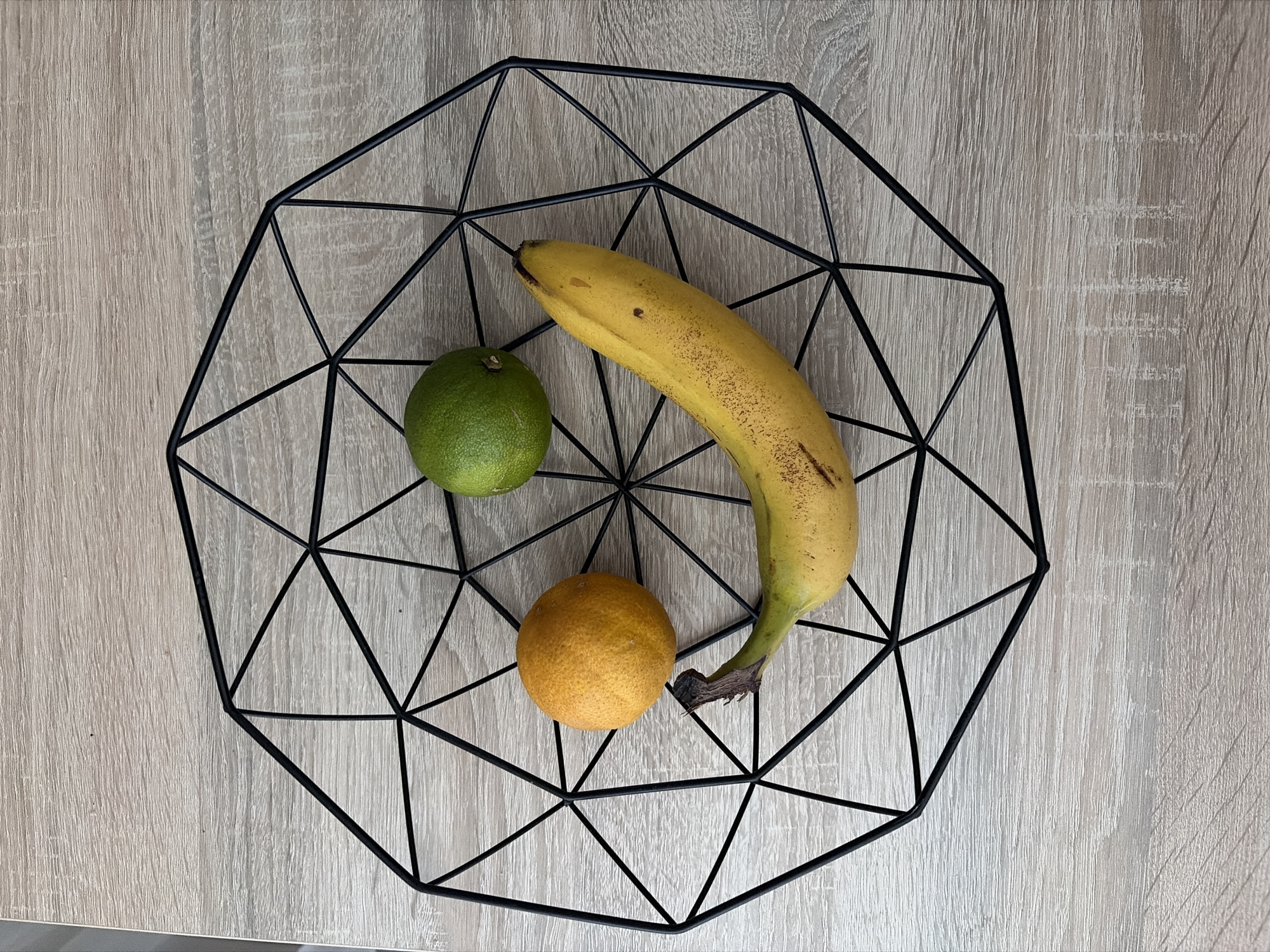 A black wire fruit basket, designed with a geometric pattern, sits on a light-colored wooden table. Inside the basket are three pieces of fruit arranged to resemble a smiley face. A yellow banana, slightly curved with brown spots, forms the smile, with its stem pointing left. To the left of the banana, a round orange citrus fruit serves as the left eye. Above and to the right of the orange, a green lime acts as the right eye. The background is a wooden surface with a visible grain pattern. The location of this scene in Nitra, Slovakia is not provided by the image.