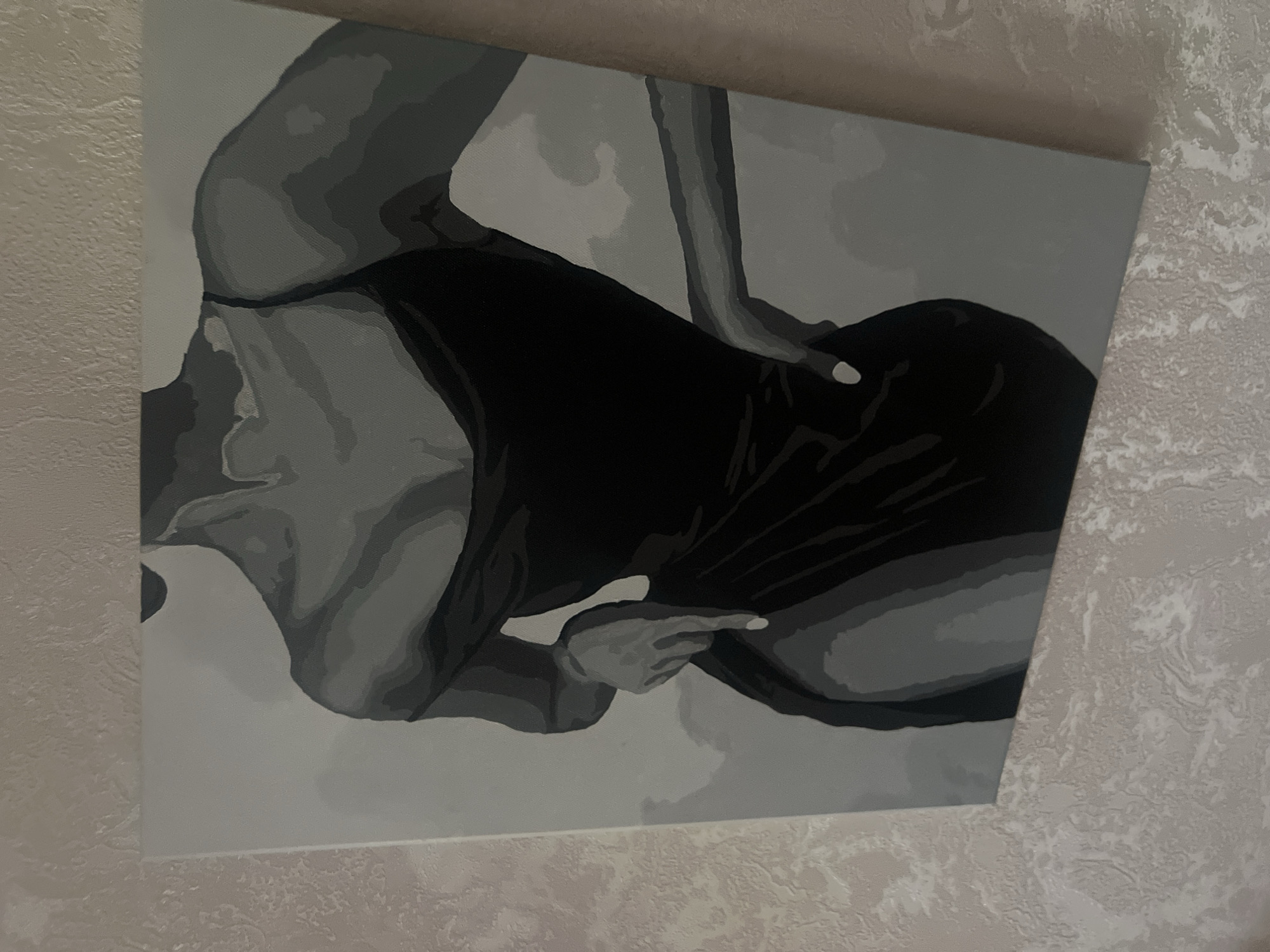 The image is a close-up, waist-up shot of a monochromatic painting of a woman in a black dress. The painting is rendered in shades of gray and black, with stark contrasts to define the form and shadows. The woman's body is angled, with her left hand placed on her hip and her right hand resting on her thigh. The dress has a thin strap and a thigh-high slit. The background of the painting is a light gray. The painting is hanging on a wall with textured wallpaper. The scene appears to be indoors, with no specific urban environmental cues.