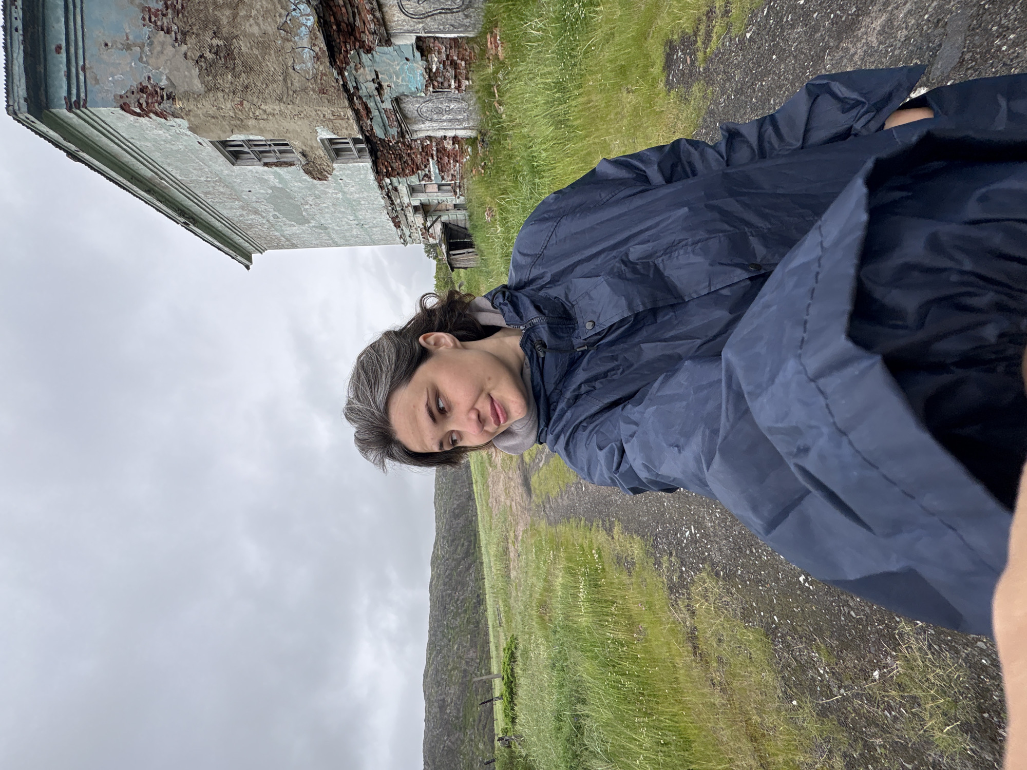 A woman with grey hair, wearing a dark blue raincoat, is standing on a dirt path in a rural area. She is looking to her left, with a slight smile. In the background, there is a green, grassy hill with a rocky texture. To the right of the path, an old, dilapidated building with peeling paint and exposed brick is visible. The sky is overcast and grey. This scene is located in Teriberka, Russia, a coastal village in Murmansk Oblast known for its stark beauty and abandoned structures.