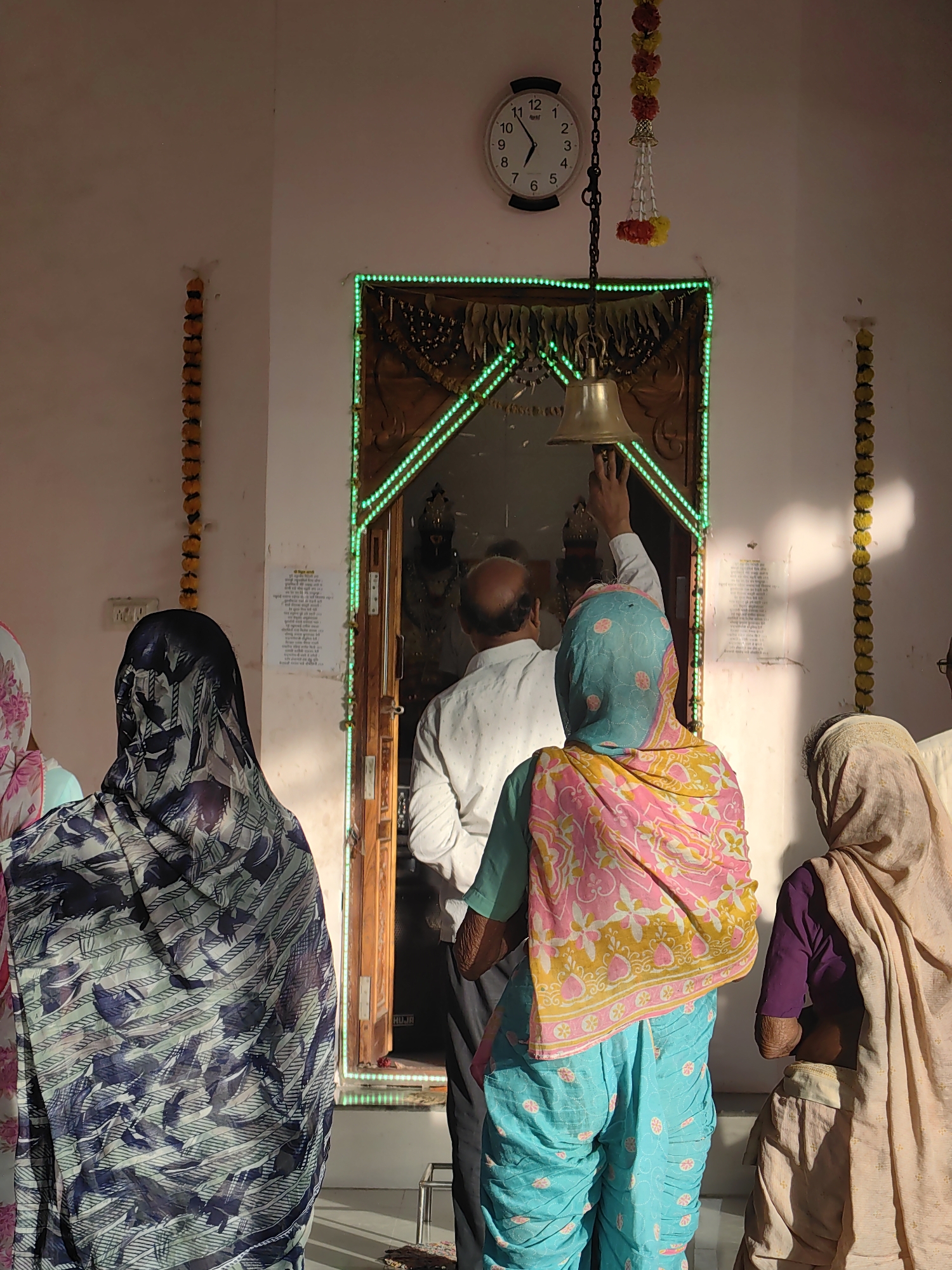 In the city of Mhaiswadi, India, a group of people are gathered outside a doorway adorned with green lights. A man in a white shirt is reaching up to ring a bell hanging above the entrance, while several women, their heads covered with colorful scarves and veils, stand in front of him. One woman wears a blue and pink patterned outfit, another a blue headscarf and pink and yellow shawl, and a third a light brown shawl. On the wall to the left, a string of orange marigold garlands hangs vertically. Above the doorway, a circular clock with black Roman numerals is mounted on the wall, showing the time to be approximately 10:57. Another string of red and yellow garlands is suspended from a chain above the bell. The doorway itself appears to be made of wood and leads into a dimly lit interior. The walls are a pale pinkish hue, and sunlight casts shadows across the scene. A bulletin board with text is visible to the right of the entrance, and to the far right, another vertical string of orange garlands is present.