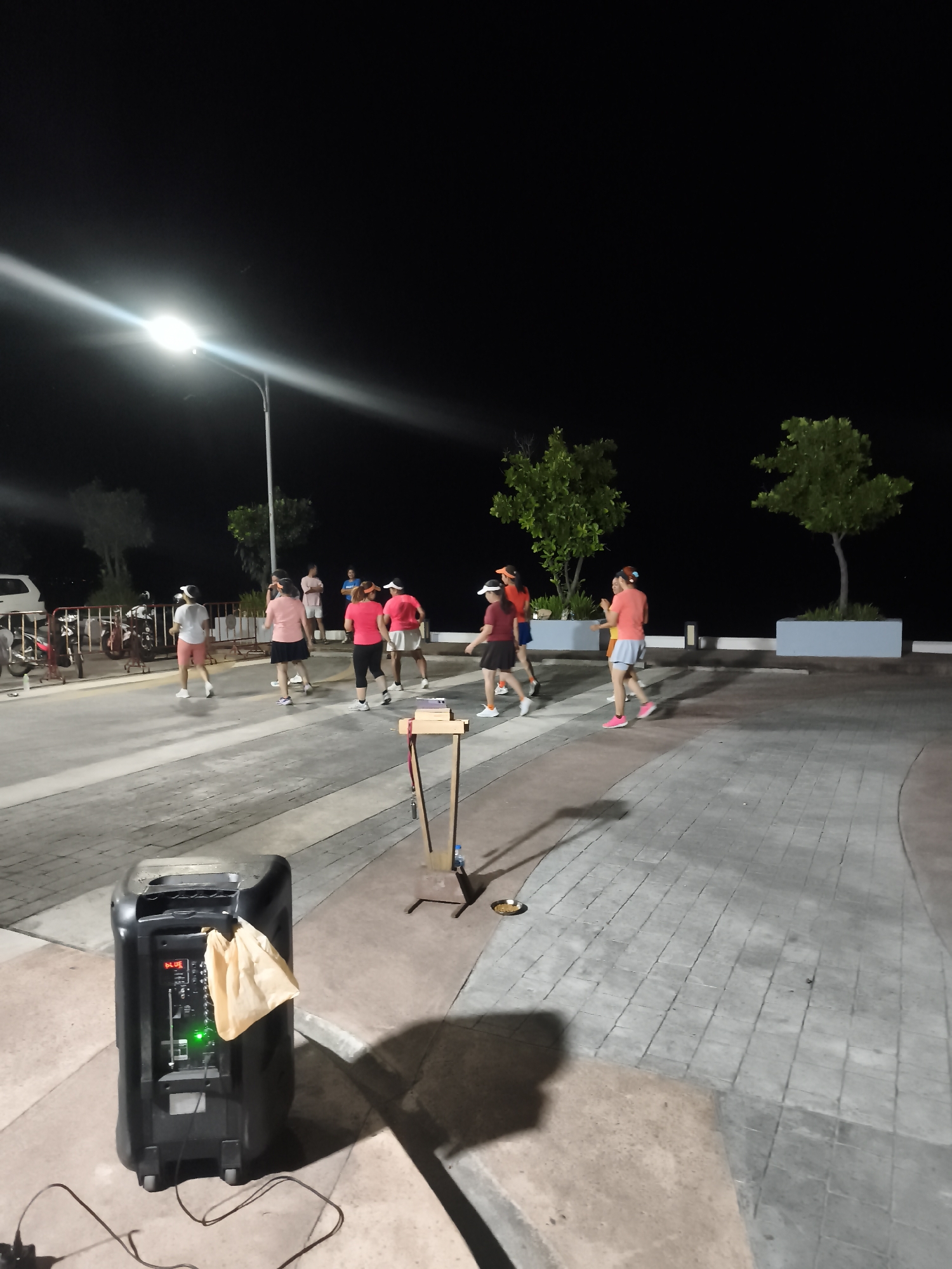 At night, a group of people participate in a social activity in Baan Hua Thanon, Thailand. The location appears to be a paved public space near a roadway with a lamppost. The participants, a group of at least 10, are engaging in what appears to be dancing or exercise, some wearing visors and light-colored shorts or skirts, and varying shades of pink and orange tops. Musical equipment, specifically a large speaker and a music stand, are set up in the foreground. The area is lined with trees in raised planters along the edges of the pavement. There are parked vehicles and motorcycles along the road.