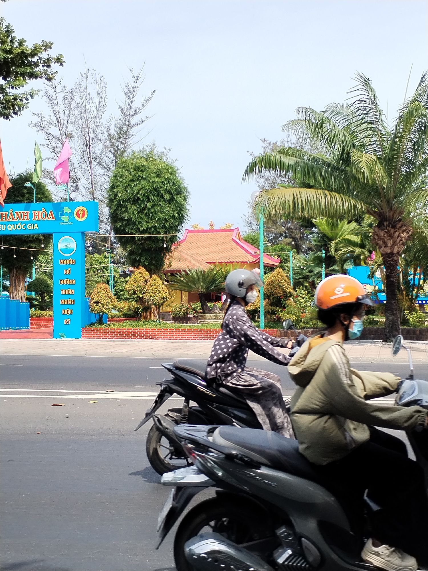 The image captures a street scene in Bac Nha Trang Ward, Vietnam, on a bright, clear day. In the foreground, two individuals are riding motorcycles. The person on the left wears a dark, star-patterned shirt and a grey helmet, their face obscured by a white mask. They are operating a dark-colored scooter. The person on the right, also wearing a mask, is on a similar scooter and is dressed in a light green hooded jacket. Their helmet is orange. Both individuals are facing forward, suggesting they are traveling along the road.
Behind the motorcycles, a blue archway stands prominent. This archway features Vietnamese text and graphics, including a circular emblem with a wave and a building illustration. The text appears to be a form of signage or advertisement. Beyond the archway, the environment transitions into a landscaped area with trees, including a tall palm tree on the right, and a building with a red, tiled roof. The ground in front of this landscaped area is lined with a low brick wall. The road itself is paved and marked with white lines. The sky is a pale blue, indicating fair weather.