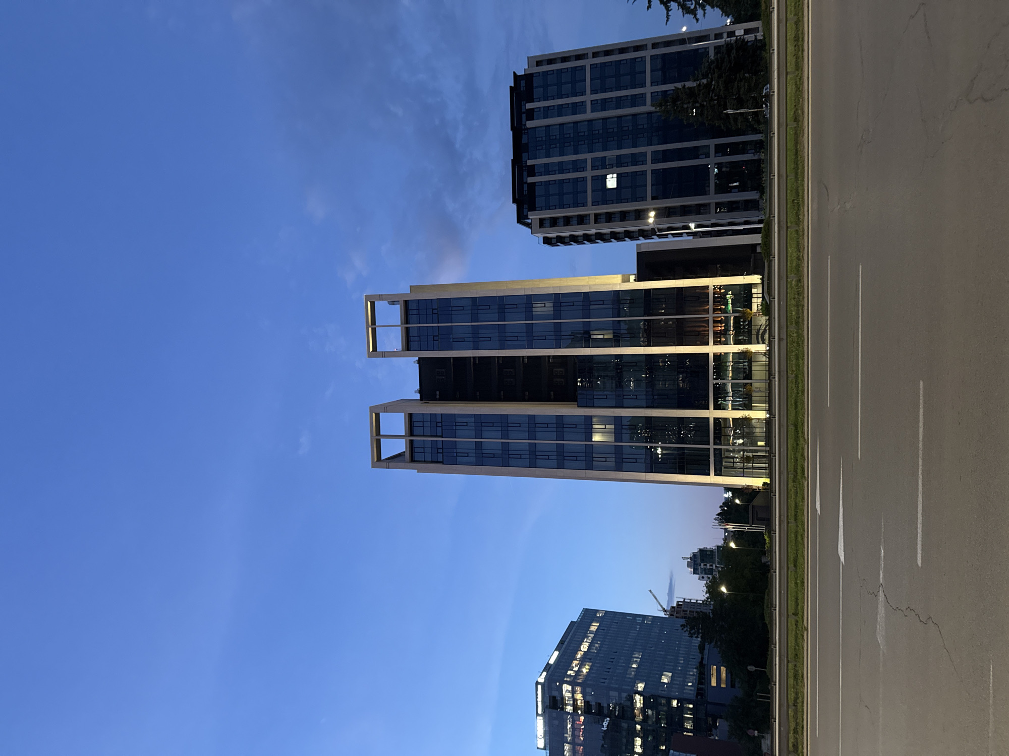 An exterior, low-angle, eye-level, full shot shows three modern office buildings on the right side of a road in Sofia, Bulgaria, at dusk. The sky is a deep blue with some wispy clouds. The building on the far left is shorter and wider than the other two, with many lit windows visible. The central building is a tall, slender skyscraper with a unique architectural design at its top, featuring a prominent, open rectangular frame. It is illuminated with vertical lights along its edges. To the right of the central building is another tall, rectangular building with dark glass windows, some of which are lit. A road with lane markings is in the foreground, and a metal guardrail separates it from a grassy area and the buildings. Trees are visible in front of the buildings. There are streetlights along the side of the road. No people are visible in the frame.