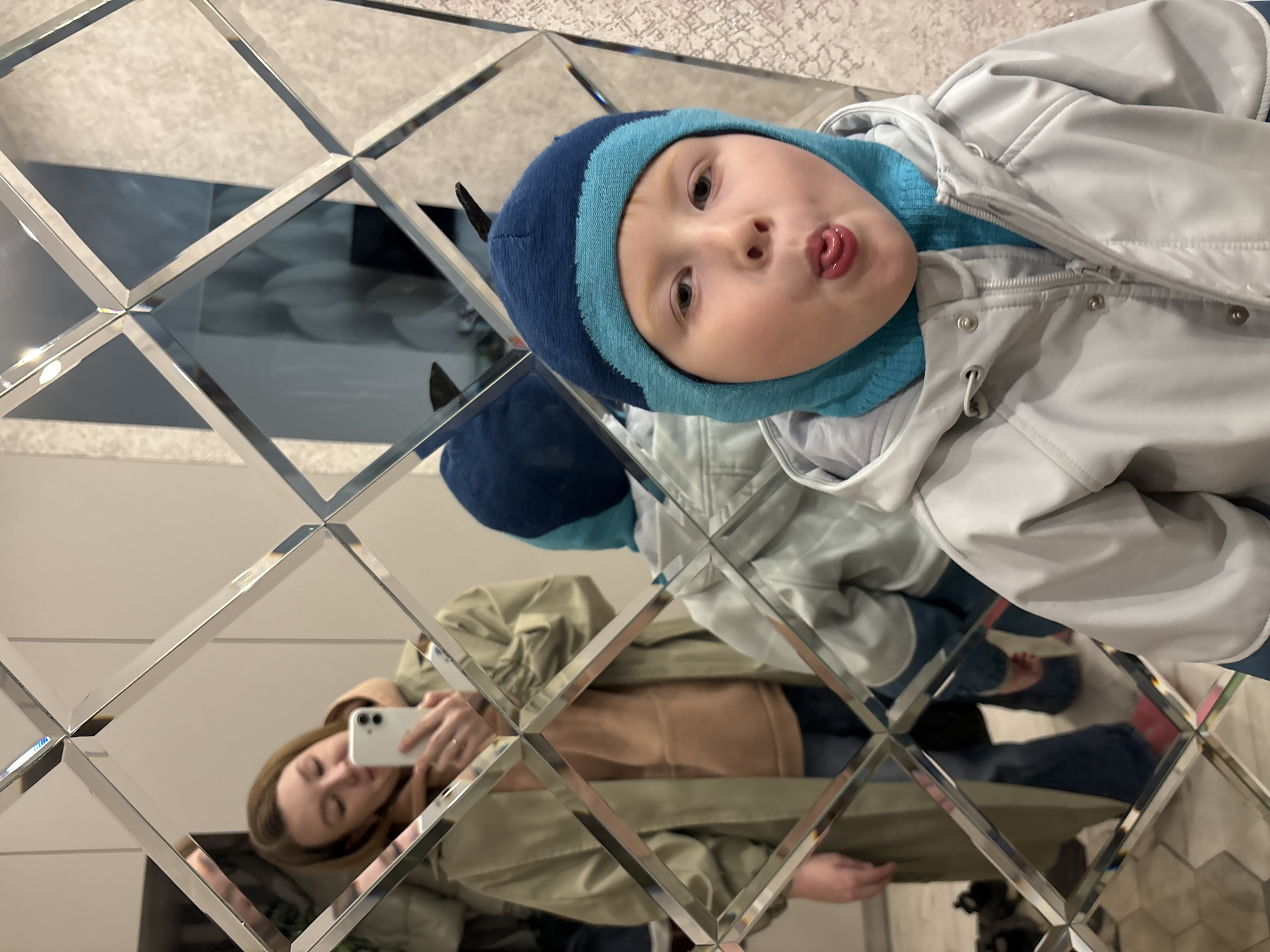A photograph taken in Izhevsk, Russia, shows a child wearing a blue and light blue striped hat and a light grey jacket making a face in a mirror made of diamond-shaped segments. A reflection in the mirror shows a woman holding a white phone, likely taking the photograph. The woman is wearing a tan jacket over a tan shirt and blue jeans. The background appears to be a wall with textured wallpaper.