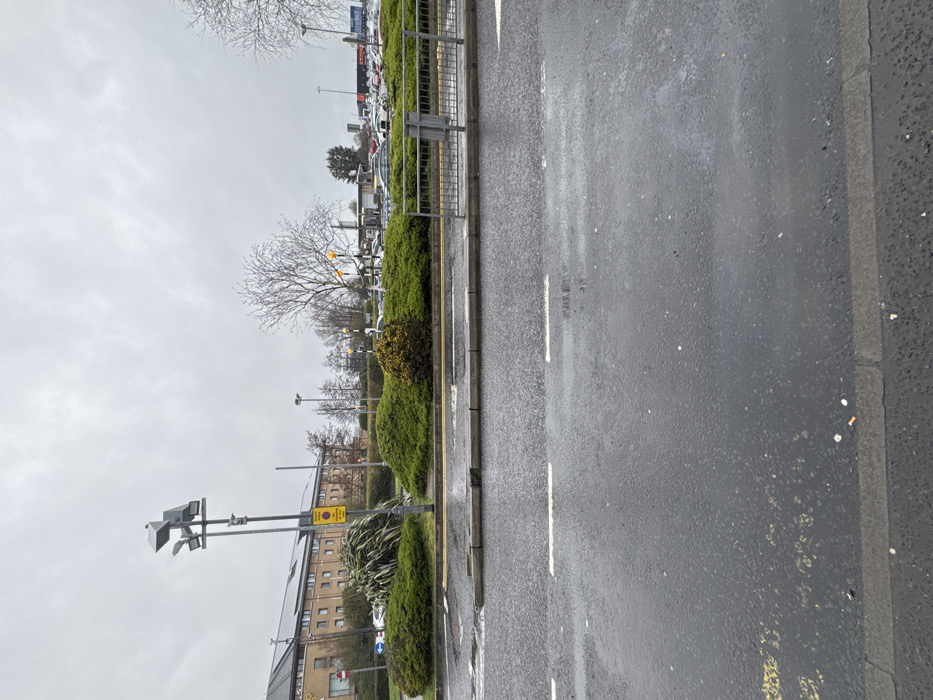 The image depicts an overcast outdoor scene in Crumlin, United Kingdom, characterized by a wet asphalt roadway and surrounding commercial infrastructure. The foreground features a dark grey asphalt road, visibly damp from rain, with two sets of broken white lane markings and faded yellow double-line markings along the curb. Scattered litter, including several cigarette butts, is present on the asphalt and the concrete curb. A low, dark grey metal pedestrian barrier extends from the right edge into the midground.
In the midground, a landscaped central reservation or roundabout is covered in dense green shrubbery, with several deciduous trees showing bare branches. A tall, dark grey utility pole with multiple light fixtures and an attached surveillance camera or sensor array stands to the left of the center. Attached to this pole are a yellow circular sign indicating "No Waiting" and a blue circular sign with a white arrow pointing left. To the left, a multi-story building with a light brown or beige facade and numerous windows is visible, identified in part as a "Premier Inn" by partial signage; its roof features a section of solar panels.
The background shows a large car park filled with various vehicles. A prominent "Tesco" supermarket sign is visible above the car park on the right side. The sky is uniformly grey and overcast. No individuals are visible, and no specific actions or interactions are depicted within the scene.