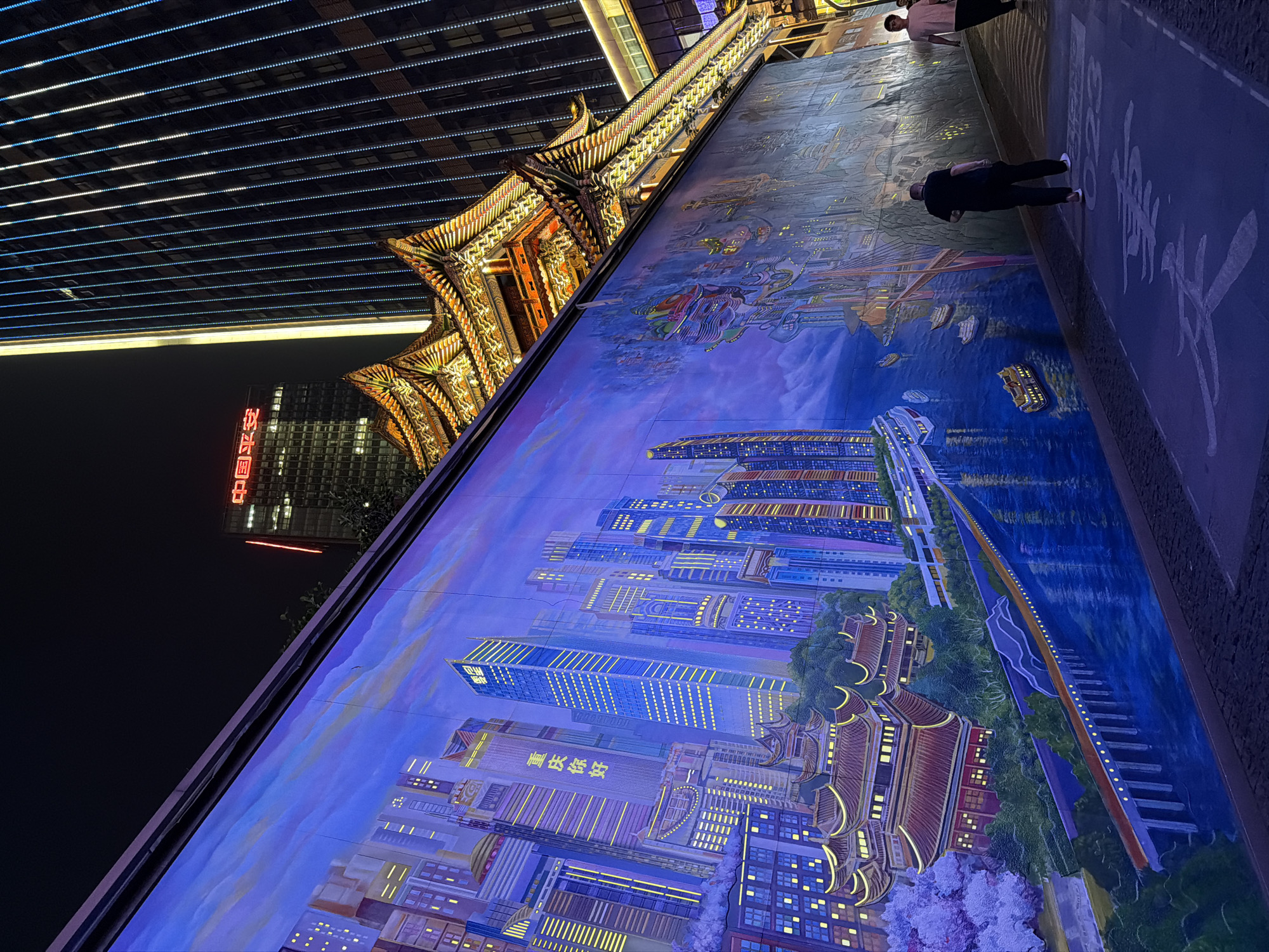 A nighttime street scene in the Yuzhong District, China, featuring a large mural depicting a cityscape, combining modern and traditional architectural styles. The mural incorporates tall, illuminated skyscrapers alongside traditional Chinese buildings with ornate roofs.
A walkway in front of the mural is visible. Two people are present: one standing and one hunched over, seemingly observing the mural. Chinese characters are painted on the walkway surface.
Above the mural and the walkway, there's a building with a traditional Chinese architectural design, illuminated with warm light. Above this building, another structure with a grid-like pattern of lights is also visible.
In the background, a taller building with illuminated Chinese characters on its side rises into the night sky.