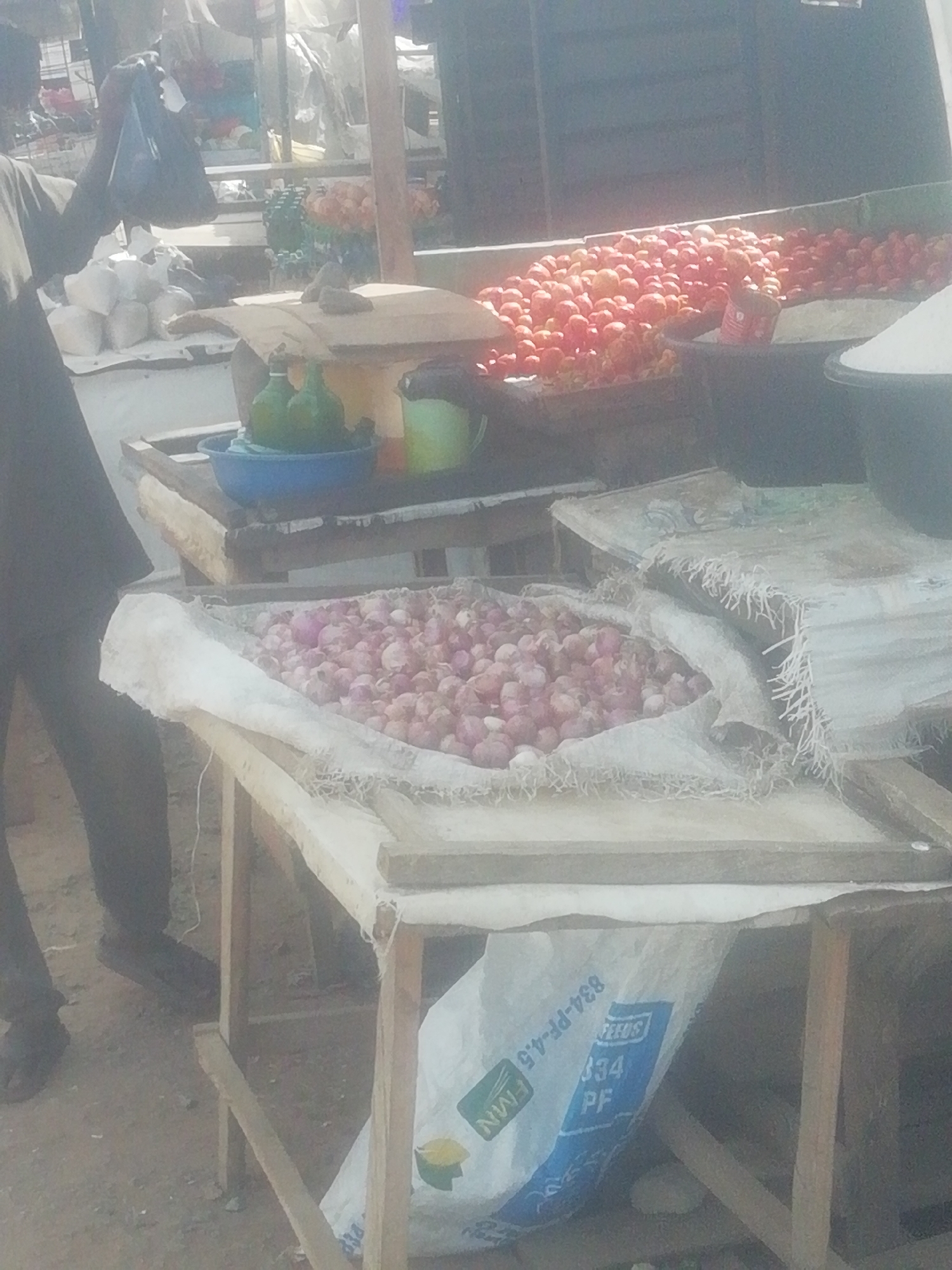 The image depicts a market scene in Ikorodu, Nigeria. A person is standing to the left, partially visible, holding a dark plastic bag. Several wooden tables display produce. One table holds a large pile of red onions in a sack. Another table has tomatoes, while a third has green bottles in a blue bowl and other items. Bags are visible beneath the tables. The environment appears to be outdoors.