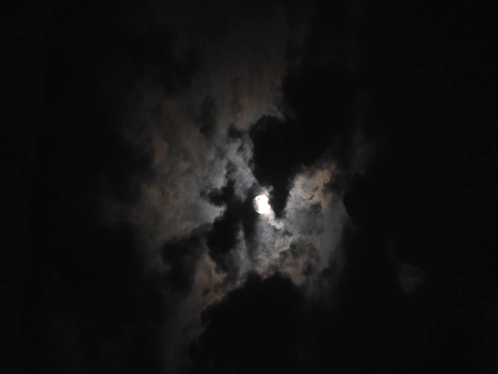 The image depicts a night sky dominated by dark, heavy clouds with a bright, full moon partially obscured by them. The moon is a luminous orb, shining through breaks in the cloud cover, creating a dramatic and atmospheric scene. The surrounding clouds are a mix of deep black and lighter grays, with some appearing almost translucent where the moonlight penetrates. The overall impression is one of a cloudy night, where the moon attempts to assert its presence through the atmospheric veil. There are no visible signs of human activity or specific landmarks that would identify a location within Ермолаево, Russia.