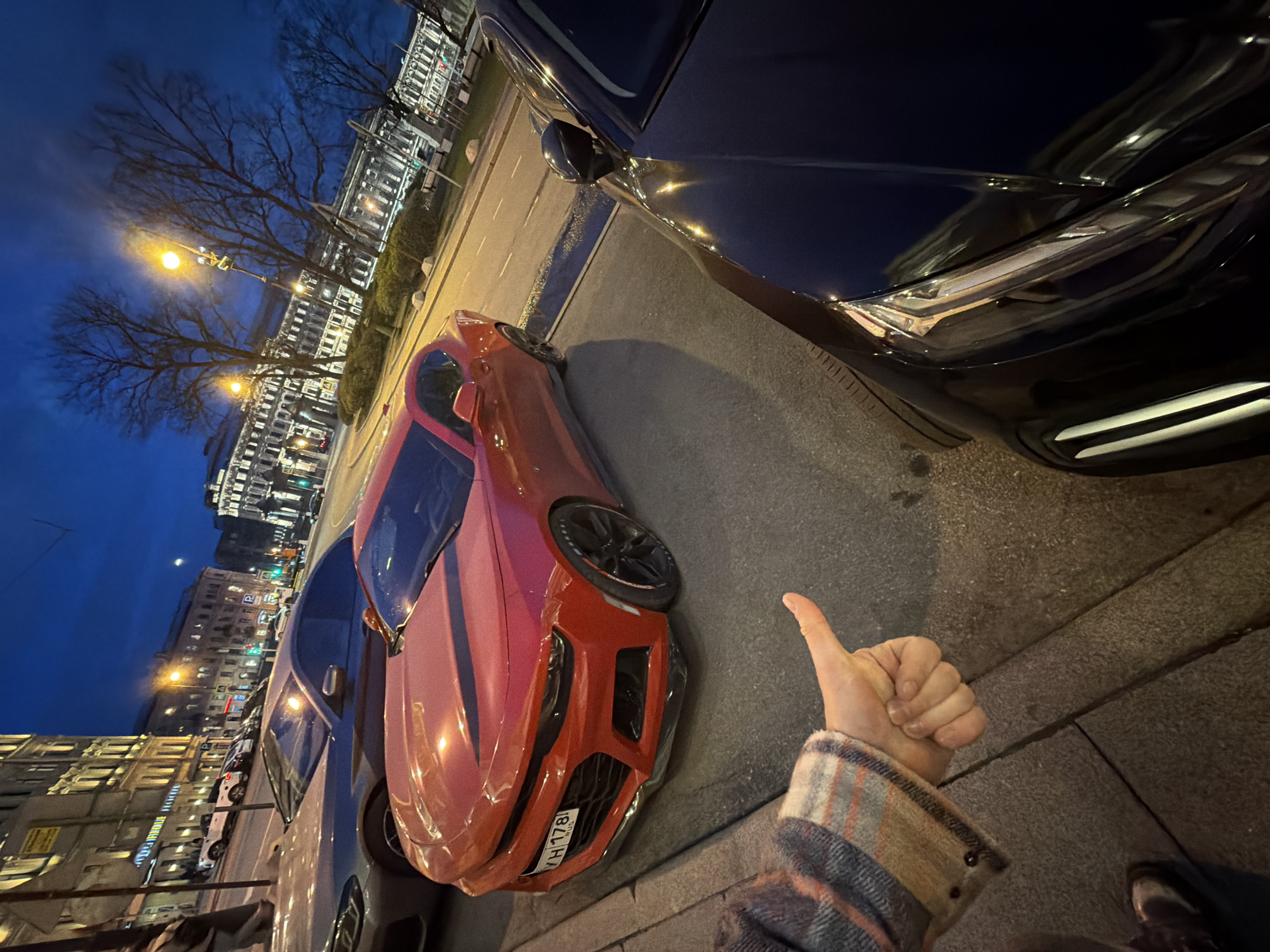 This media file captures an evening or night street scene in Saint Petersburg, Russia. The image is taken from a low angle, looking down a street lined with buildings and parked cars.
In the immediate foreground, a person's left hand is visible, giving a "thumbs up" gesture. The hand wears a sleeve of a plaid, multi-colored jacket (beige, blue, orange).
The primary subject is a vibrant red sports car, likely a Chevrolet Camaro, parked parallel to the curb. Its front end, including the grille and headlights, is clearly visible, facing towards the right of the frame. A Russian license plate, "YH 178 | RUS", is attached to its front bumper. To the right of the red car, a dark blue or black car is partially visible, showing its side profile and highly reflective surface, mirroring the streetlights and surrounding environment. Other cars are parked along the street further down.
The background features classic European-style multi-story buildings, illuminated by their interior lights, suggesting residential or commercial use. Bare-branched trees line the street, indicating a cold season (late autumn, winter, or early spring). Several streetlights cast a warm yellow glow, illuminating the street and sidewalk. The sky is a deep, dark blue, characteristic of twilight or night. A faint, bright light (possibly the moon or another distant light source) is visible in the sky.
The overall scene depicts an urban setting after dusk, with the thumbs-up gesture possibly expressing approval or appreciation for the red sports car.