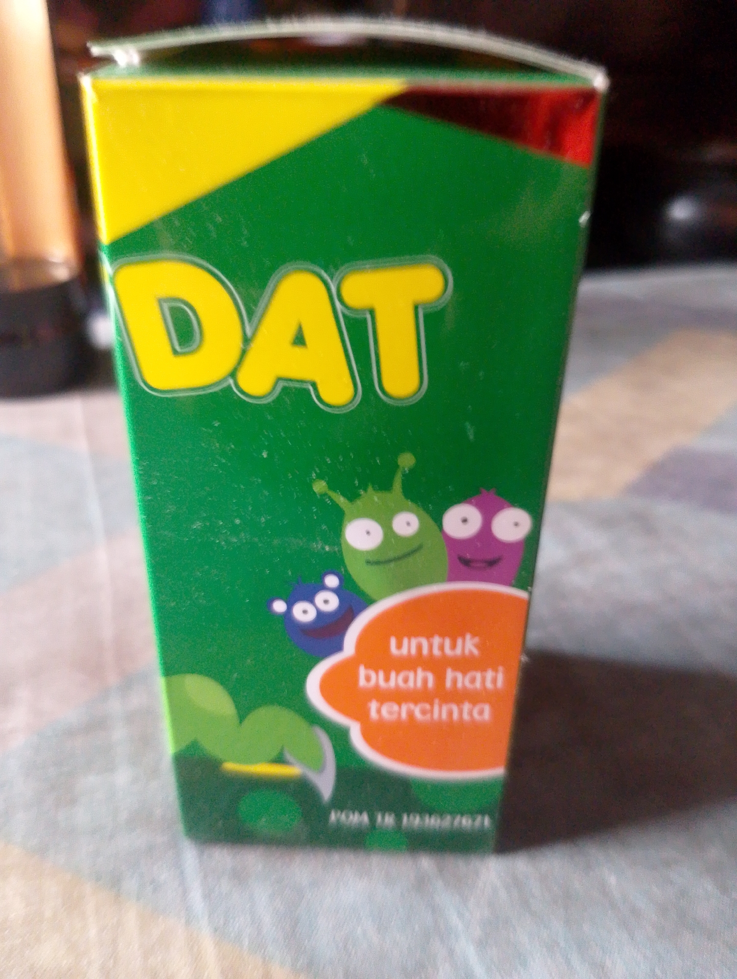 The image shows a green and yellow box with the word "DAT" in large yellow letters. There are cartoonish characters on the box, along with text in Indonesian that says "untuk buah hati tercinta" which translates to "for beloved fruit of the heart." At the bottom of the box, there is text that appears to be a product registration number. The box is sitting on a patterned surface, possibly a tablecloth, and is partially lit by a warm light source, suggesting an indoor setting. The background is blurred and out of focus, but appears to be a domestic environment. There is no indication of the city of South Jakarta, Indonesia in the image.
