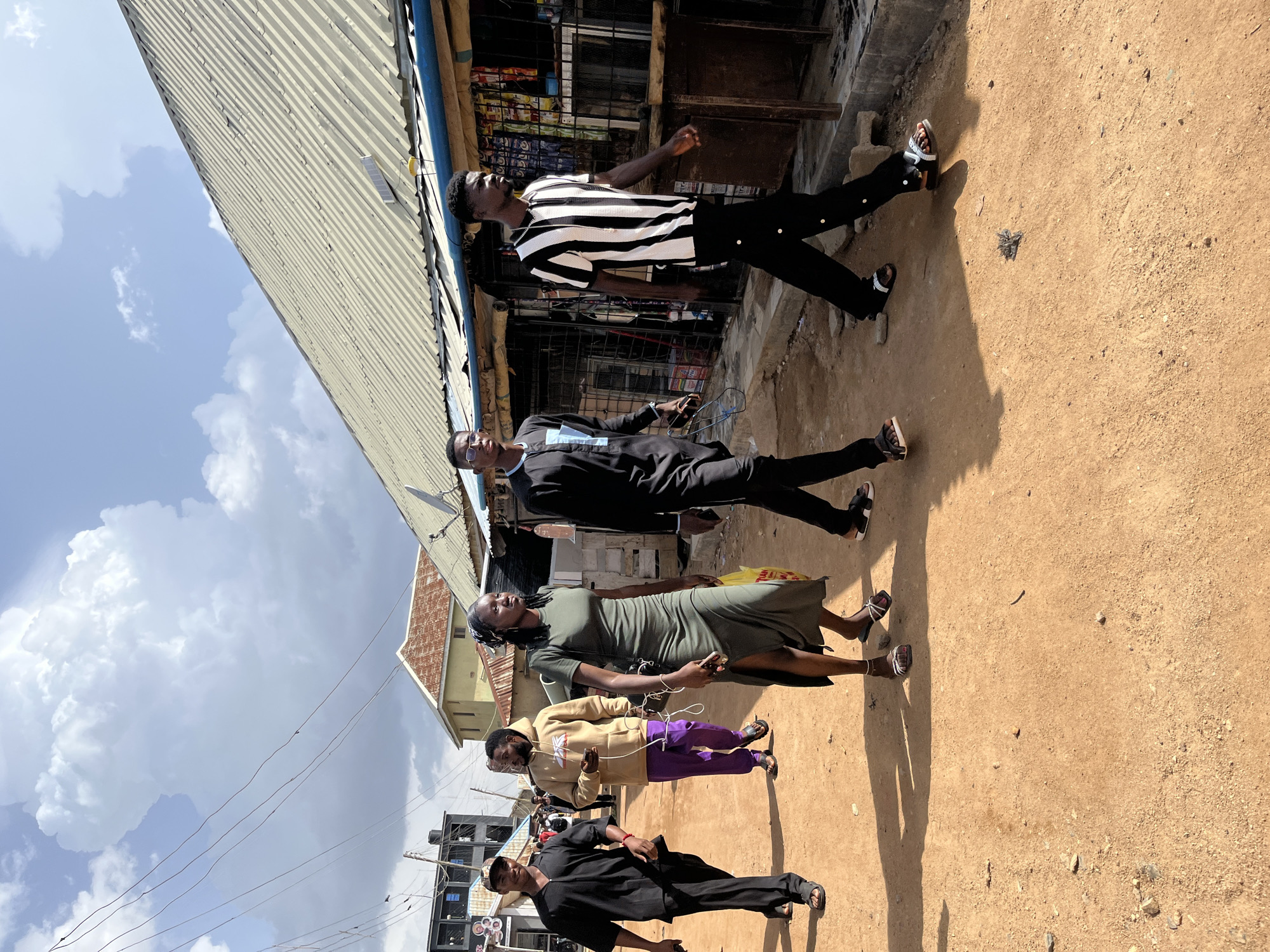 In Jos, Nigeria, a group of five young adults walk down a dusty, unpaved street under a partly cloudy sky. The individuals, four men and one woman, are walking from left to right in the frame. The man on the far left wears a black V-neck tunic and trousers, with a dark baseball cap. Next to him, a man in a tan hoodie and purple pants holds a mobile phone. The woman in the center wears a long, olive-green dress with a high slit and carries a yellow plastic bag. She is followed by two men; one in a black and blue patterned tunic, and the other in a black and white striped shirt and black trousers. They are walking past a row of shops with corrugated metal roofs, displaying various goods for sale. The ground is a reddish-brown dirt, with scattered stones and debris. The buildings in the background appear to be a mix of residential and commercial structures, with some showing signs of wear. Power lines crisscross the sky overhead.
