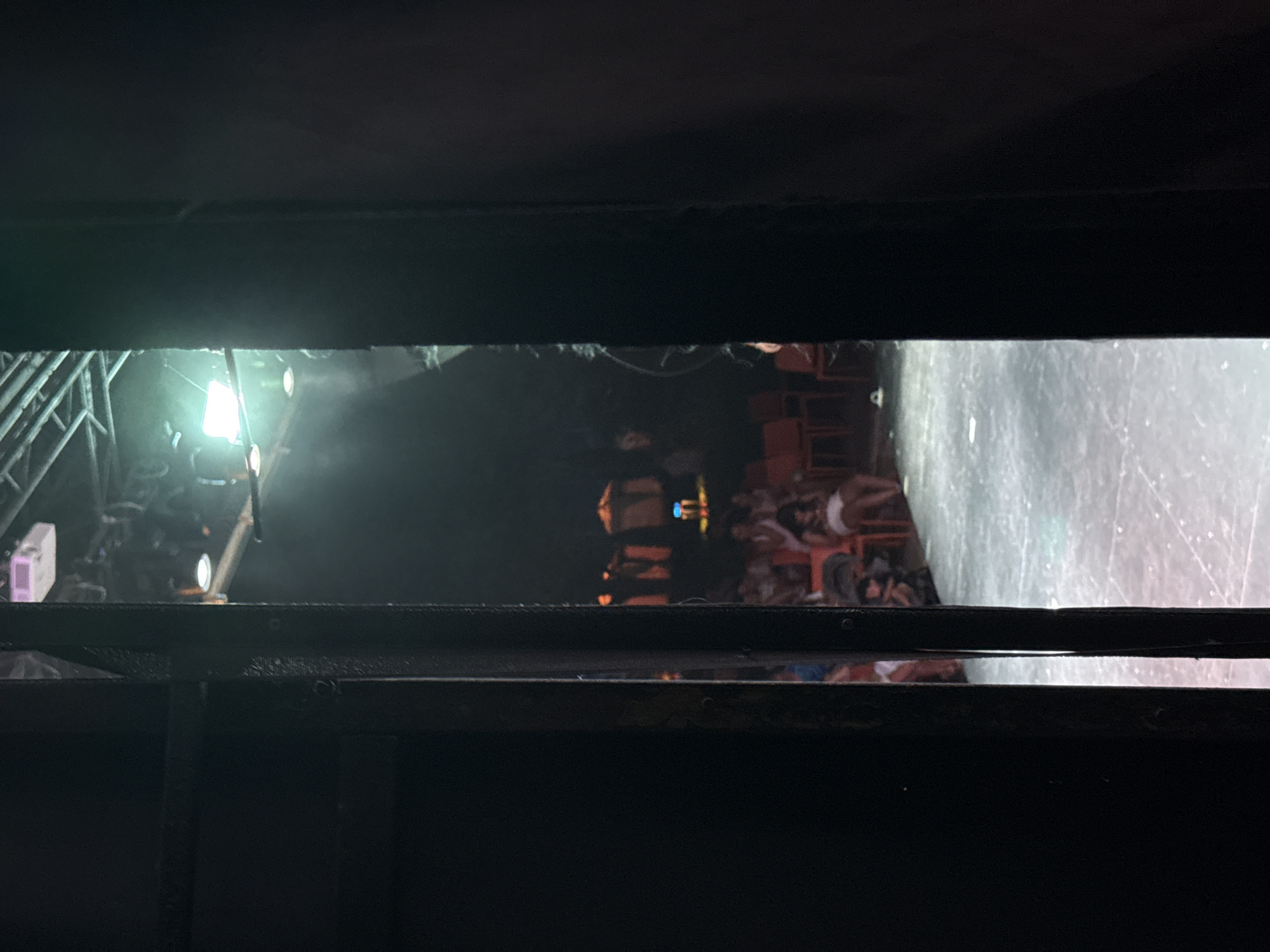 This is a photograph taken from behind a black frame, looking out into a dimly lit theater or performance space. The frame is positioned to the left of the image and a darker, narrower frame is to the right, creating a view through a narrow opening.
In the background, a stage area is visible with a light-colored floor that appears to be speckled or textured. Scattered across the floor are what look like small white debris or marks. Upstage, rows of orange and red chairs are occupied by an audience. The people are mostly indistinct due to the lighting and distance, but some appear to be wearing light-colored clothing, possibly white or light blue. Some individuals are seated, while others might be standing or in the process of sitting.
Above the stage, stage lighting equipment is visible, including what appears to be a projector on the ceiling and several spotlights. One bright, square light source is prominent, casting a green glow on the scene. The overall atmosphere is dark and moody, suggesting a performance is about to begin or has recently concluded.
There is no indication of the city of Aksu, Turkey, within the image itself. The description is limited to the visual information presented in the photograph.