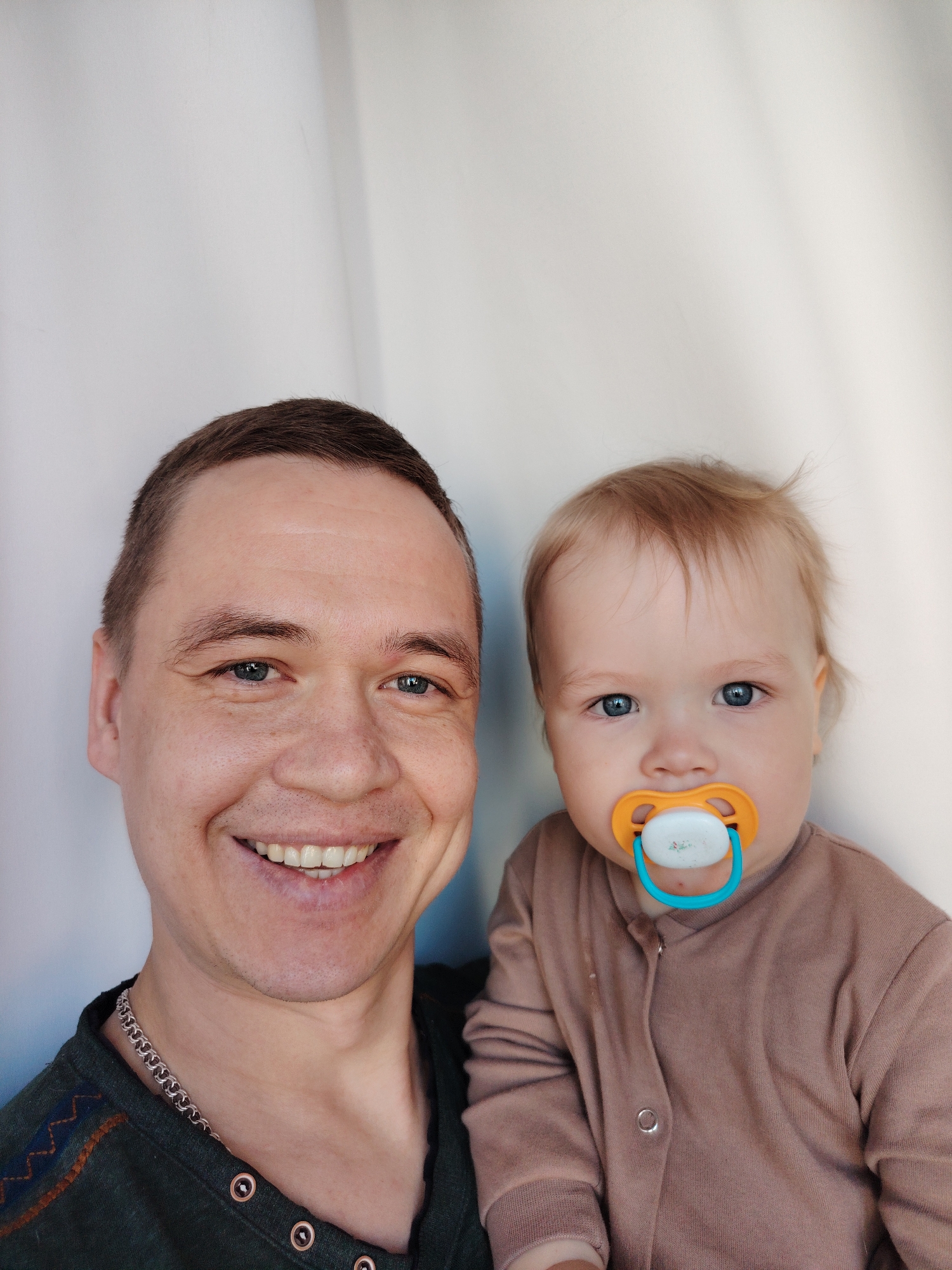 A man and a baby are pictured in a close-up selfie. The man is on the left with short, dark hair, light skin, and blue eyes, smiling broadly, revealing his teeth. He is wearing a dark green shirt with decorative stitching. The baby, on the right, has fair skin, light blonde hair, blue eyes, and a pacifier in their mouth. The baby is wearing a brown onesie. The background is a plain, off-white or light grey wall with subtle shadows. There is no indication of location or the city of Chaikovsky, Russia, in the image.