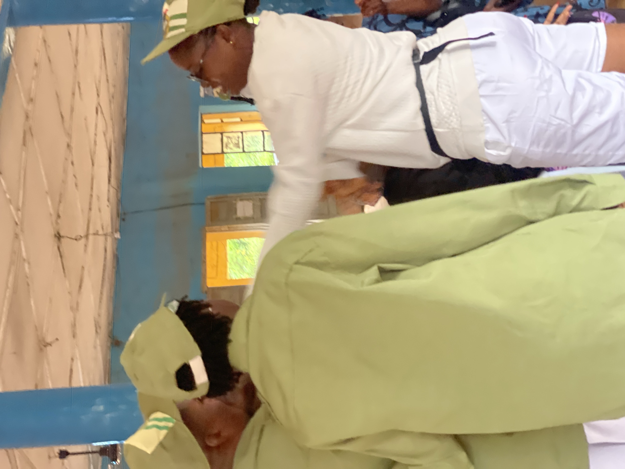 Three individuals are visible in the foreground, two wearing light green shirts and matching caps, and one wearing a white long-sleeved shirt and white shorts. The person in white is facing away from the viewer, with their right arm extended towards the person on the left in the green shirt. The person on the left in the green shirt is also facing away from the viewer, their left shoulder visible. The third individual, also in a green shirt and cap, is partially obscured between the other two. The environment appears to be an indoor space with a light-colored, textured ceiling and blue walls. Two windows with orange frames are visible in the background, through which greenery can be seen. The scene takes place in Ise-Ekiti, Nigeria.