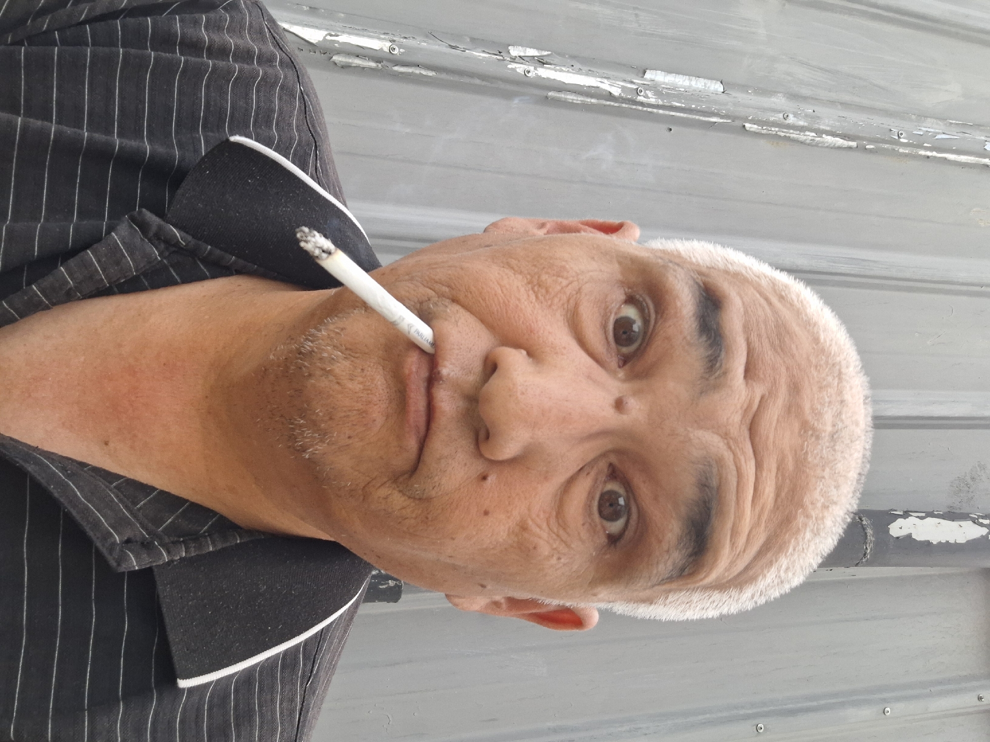 This is a close-up, portrait-style image of a man with a cigarette in his mouth. He has short, light-colored hair and is wearing a black collared shirt with thin white pinstripes. His eyes are open, and he appears to be looking directly at the camera with a neutral or slightly surprised expression. The background is a corrugated metal surface, which is weathered and shows signs of peeling paint in places. The lighting suggests it is daytime, possibly outdoors, given the texture and appearance of the background. The cigarette is lit and smoking. There is no discernible text in the image.