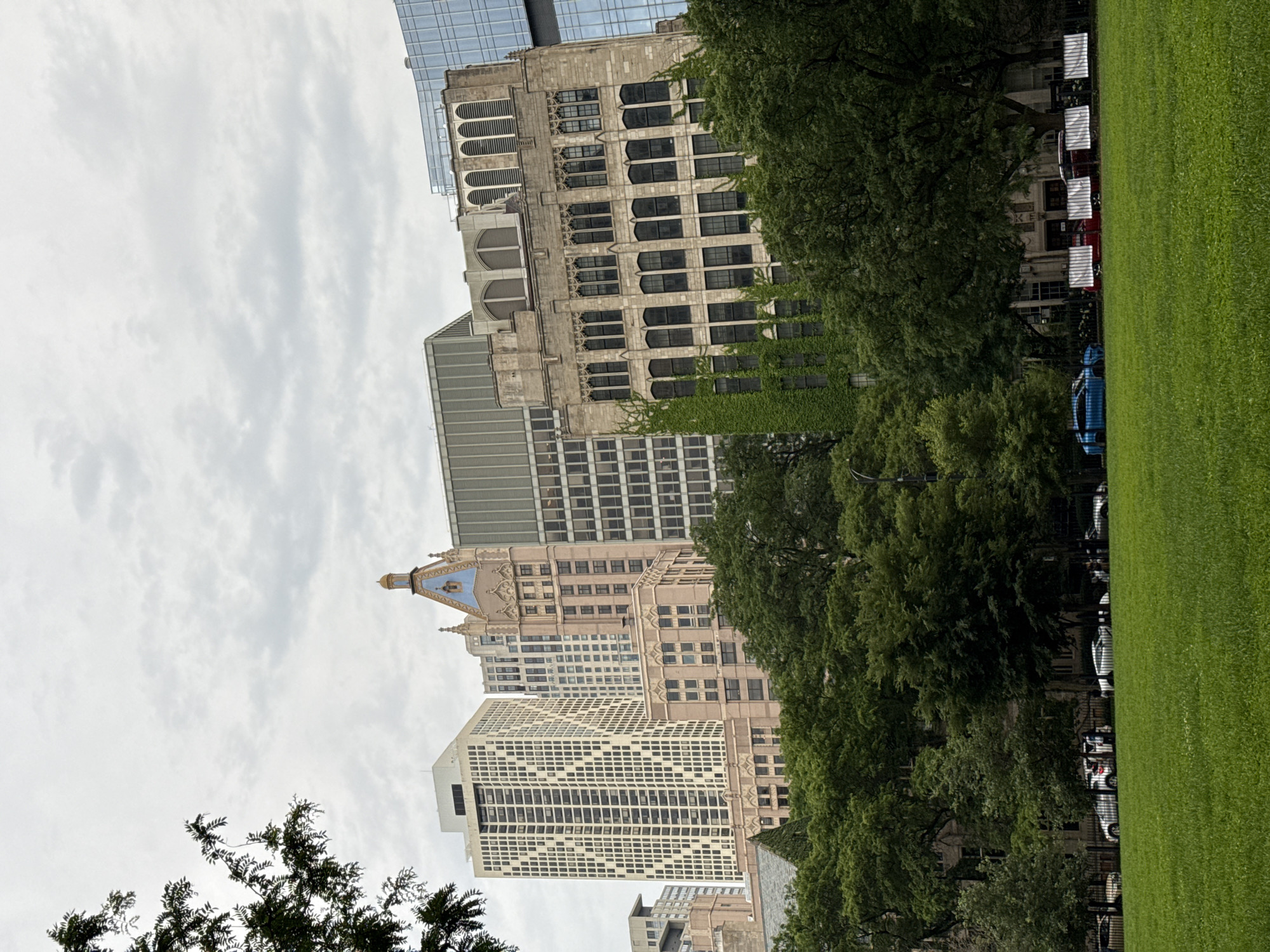 This image depicts a city park in Chicago, Illinois, with a view of several tall buildings in the background. In the foreground, a lush green lawn stretches across the scene, with mature trees lining the edge of the park and partially obscuring the buildings. A black metal fence separates the lawn from a sidewalk where several parked cars are visible. Beyond the trees, various architectural styles of buildings are present, including a tall, cream-colored high-rise with a geometric facade, a lighter-colored building with ornate details and a blue spire, and a stone building covered in ivy. The sky is overcast with gray clouds.